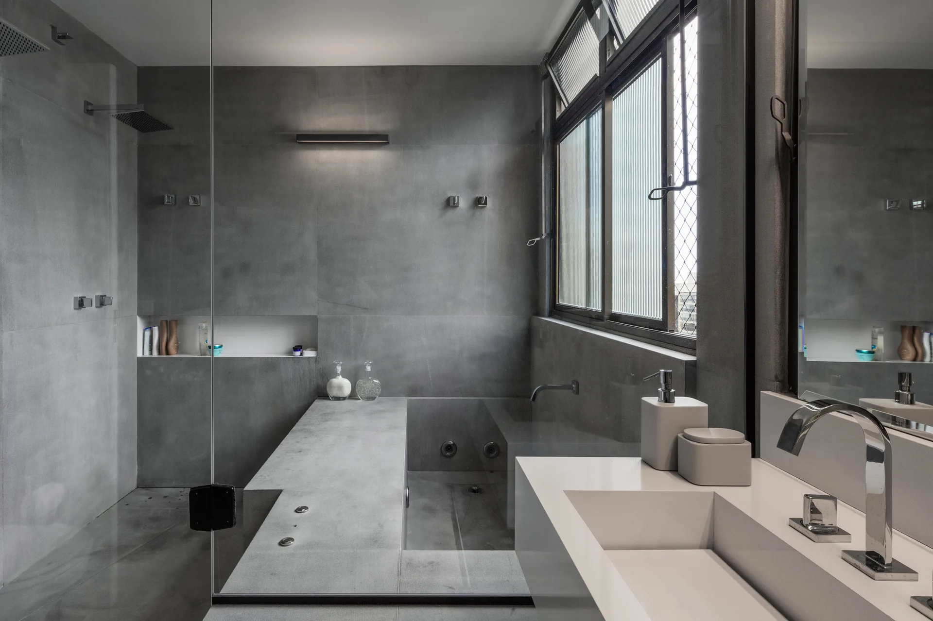 A modern gray concrete bathroom featuring a sunken soaking tub, a frameless glass shower panel, and a white integrated sink vanity.
