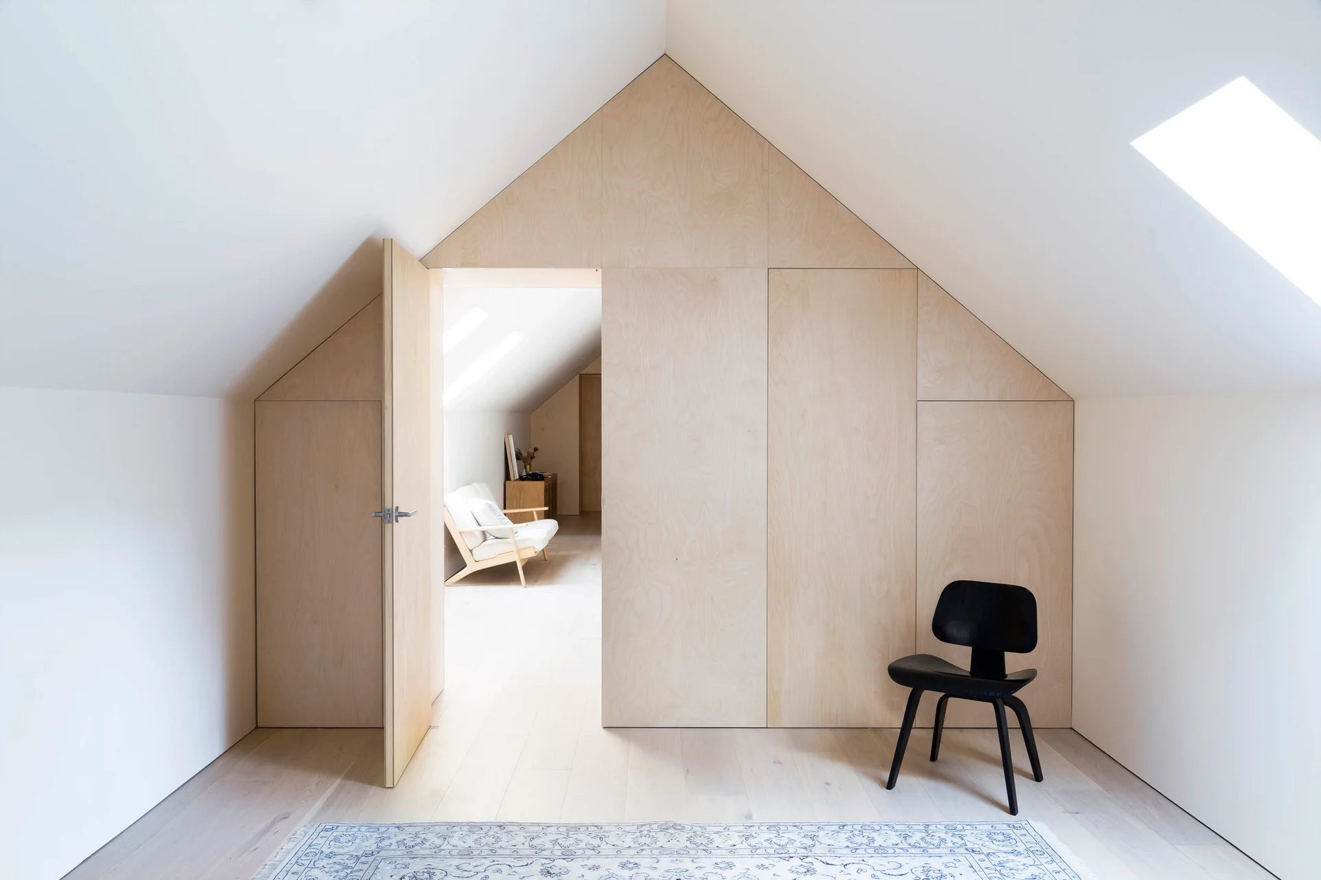 A minimalist attic bedroom features a gabled plywood storage wall with an open doorway, white sloped ceilings, and a black accent chair.