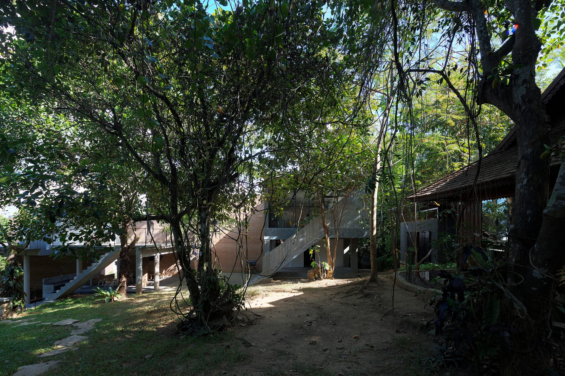Modern concrete and brick buildings with outdoor staircases are nestled within a dense forest of mature trees.