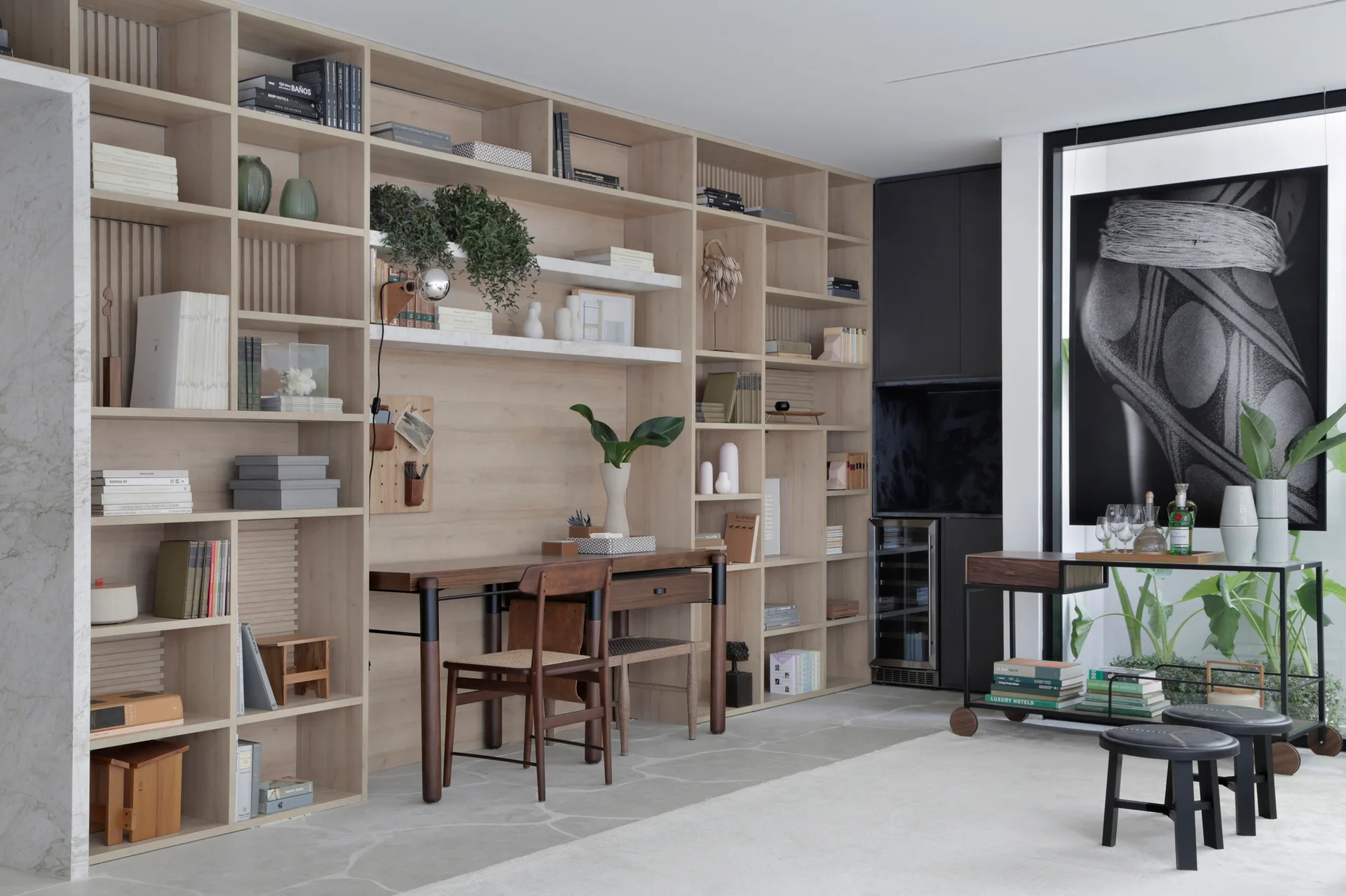 A home office features a large light wood built-in shelving unit with an integrated desk, a dark wood chair, and a rolling bar cart.