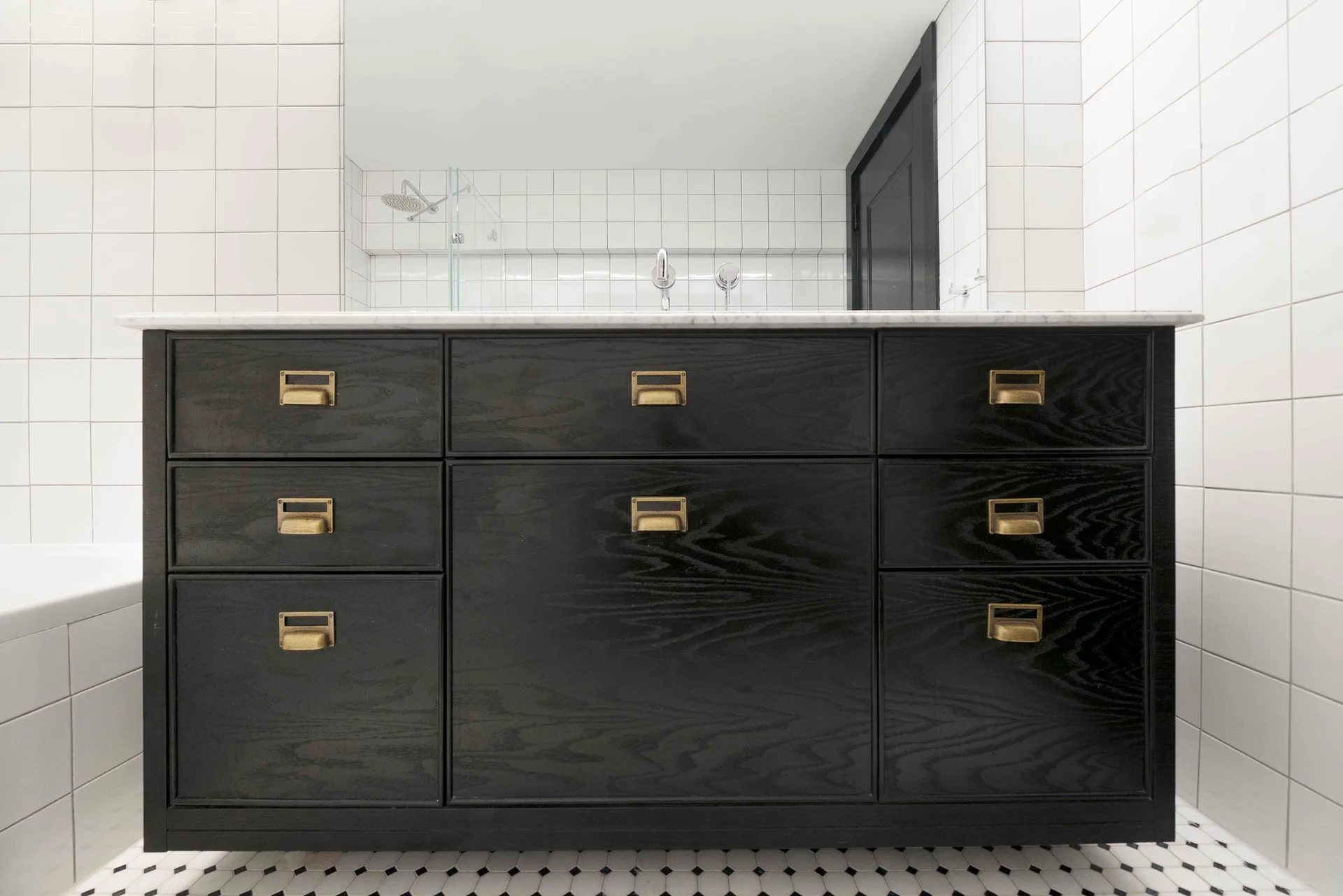 A black stained wood vanity with brass hardware sits against a white square-tiled wall with small black and white floor tiles.