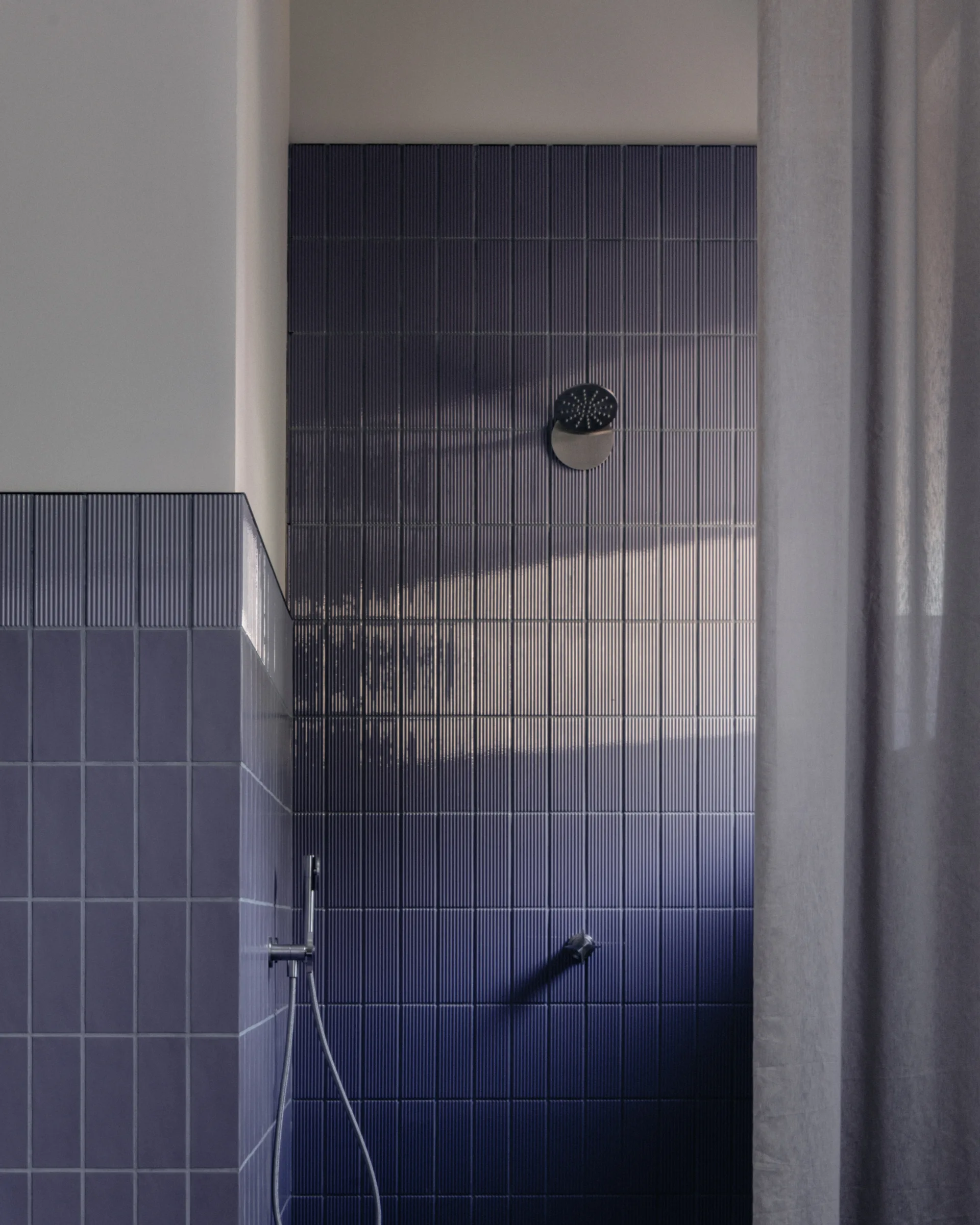 A minimalist shower features a dark blue tiled wall with vertical ribbed textures, a silver shower head, and a soft grey curtain.