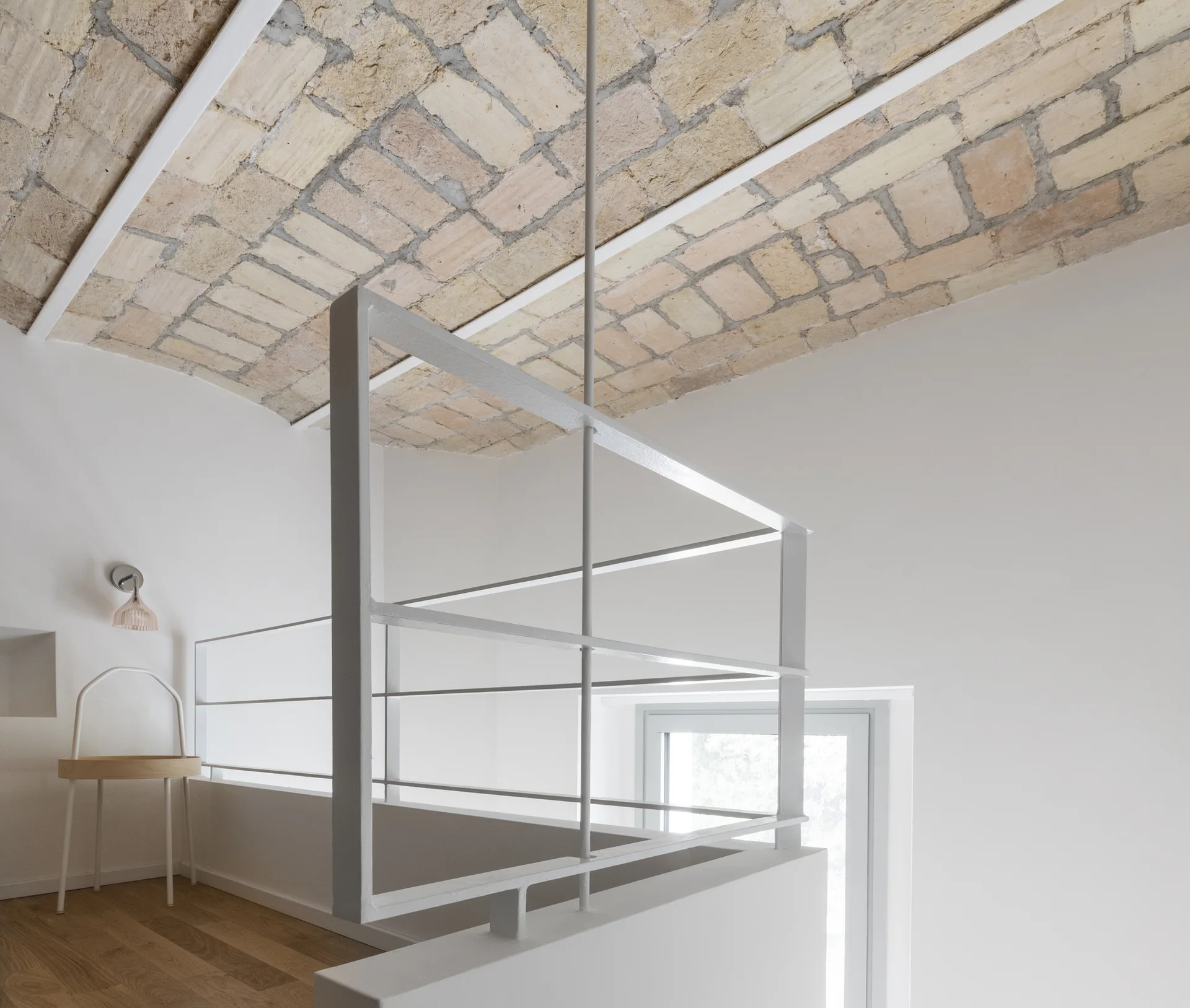 A minimalist white loft landing features an exposed brick vaulted ceiling, a thin white metal railing, and light wood flooring.