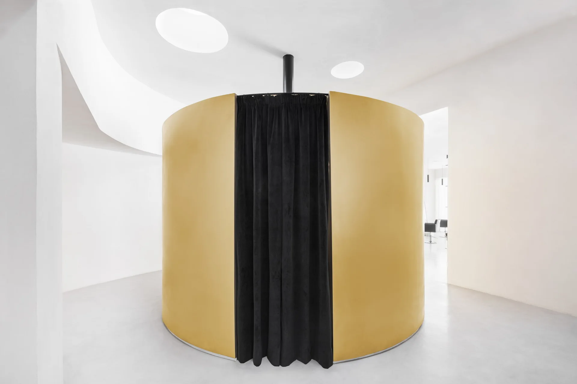 A cylindrical matte gold partition with a black fabric curtain stands in the center of a minimalist white room with circular ceiling skylights.
