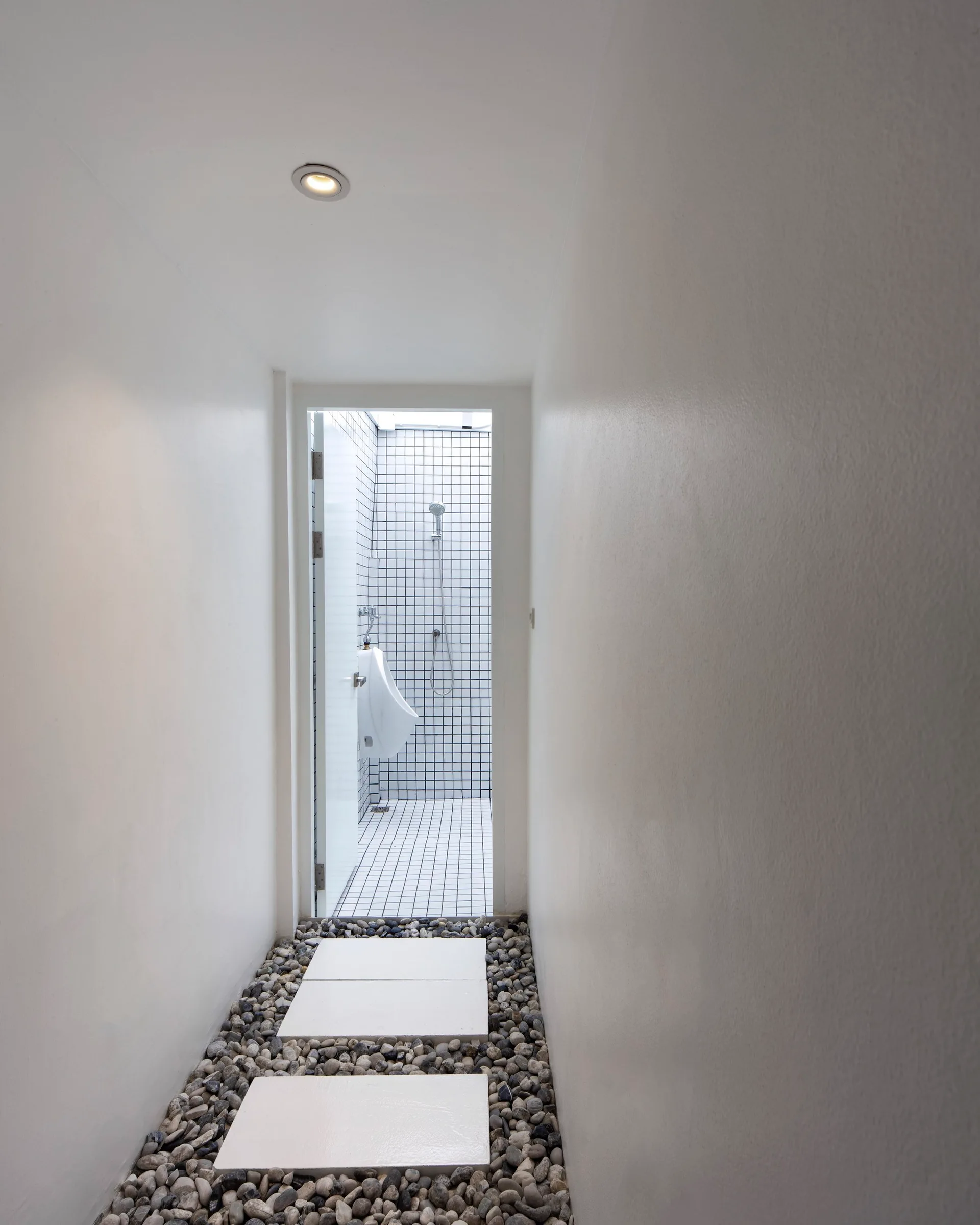 A narrow white hallway with a stone pebble pathway and stepping stones leads to a brightly lit bathroom with square grid tiles.
