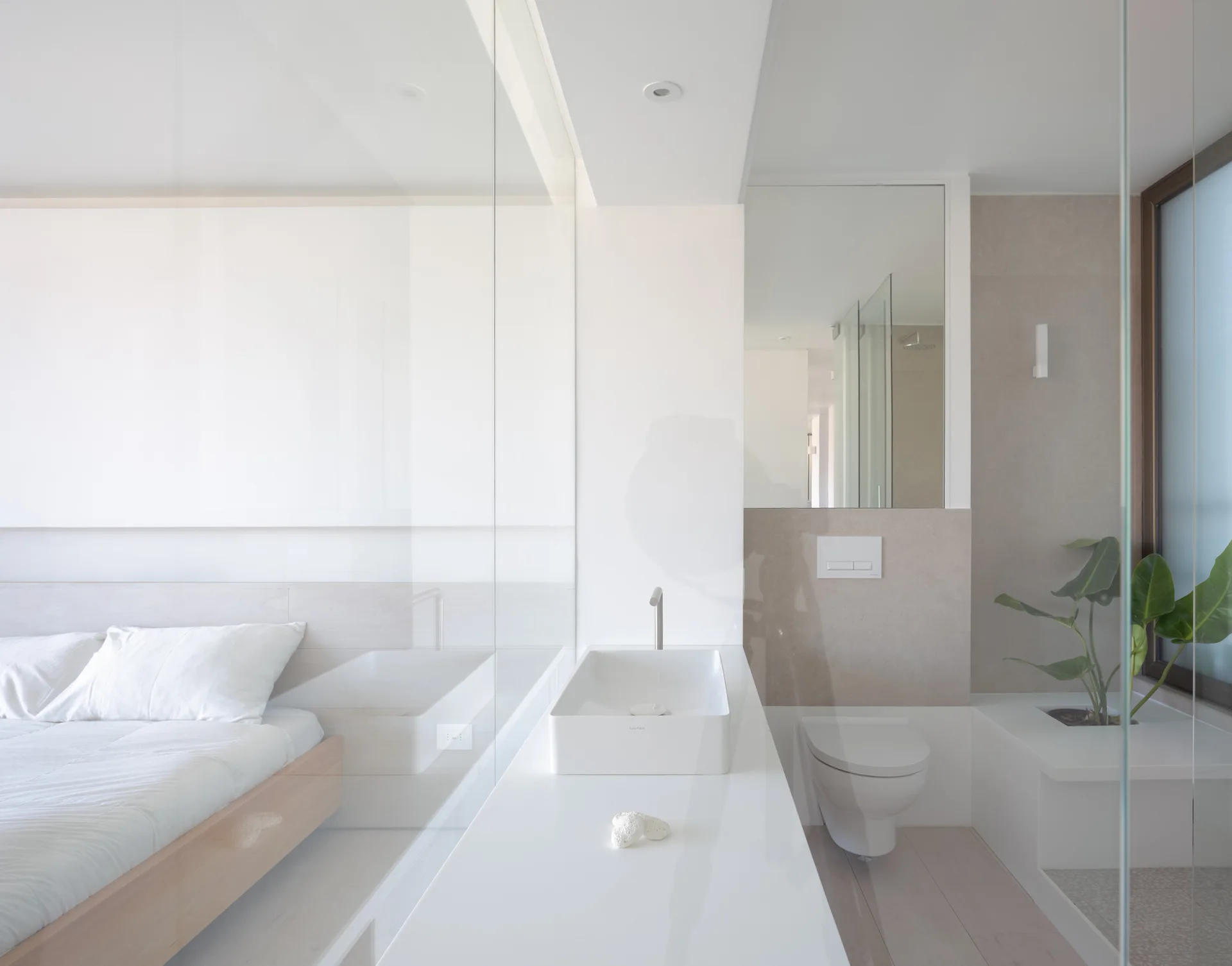 A minimalist bedroom and bathroom suite separated by a clear glass wall, featuring a low wooden bed, white countertop sink, and wall-hung toilet.