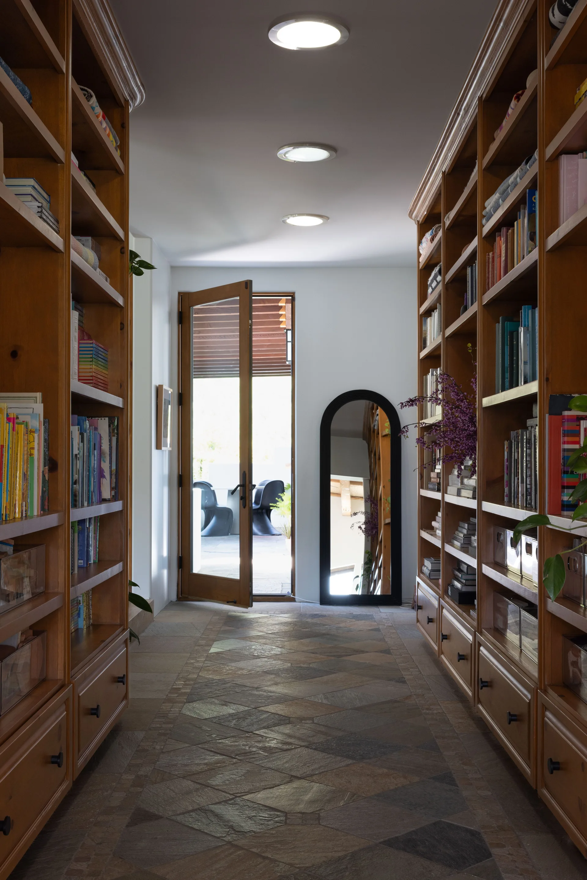 A symmetrical hallway lined with wooden built-in bookshelves features a patterned stone tile floor and an arched floor mirror.