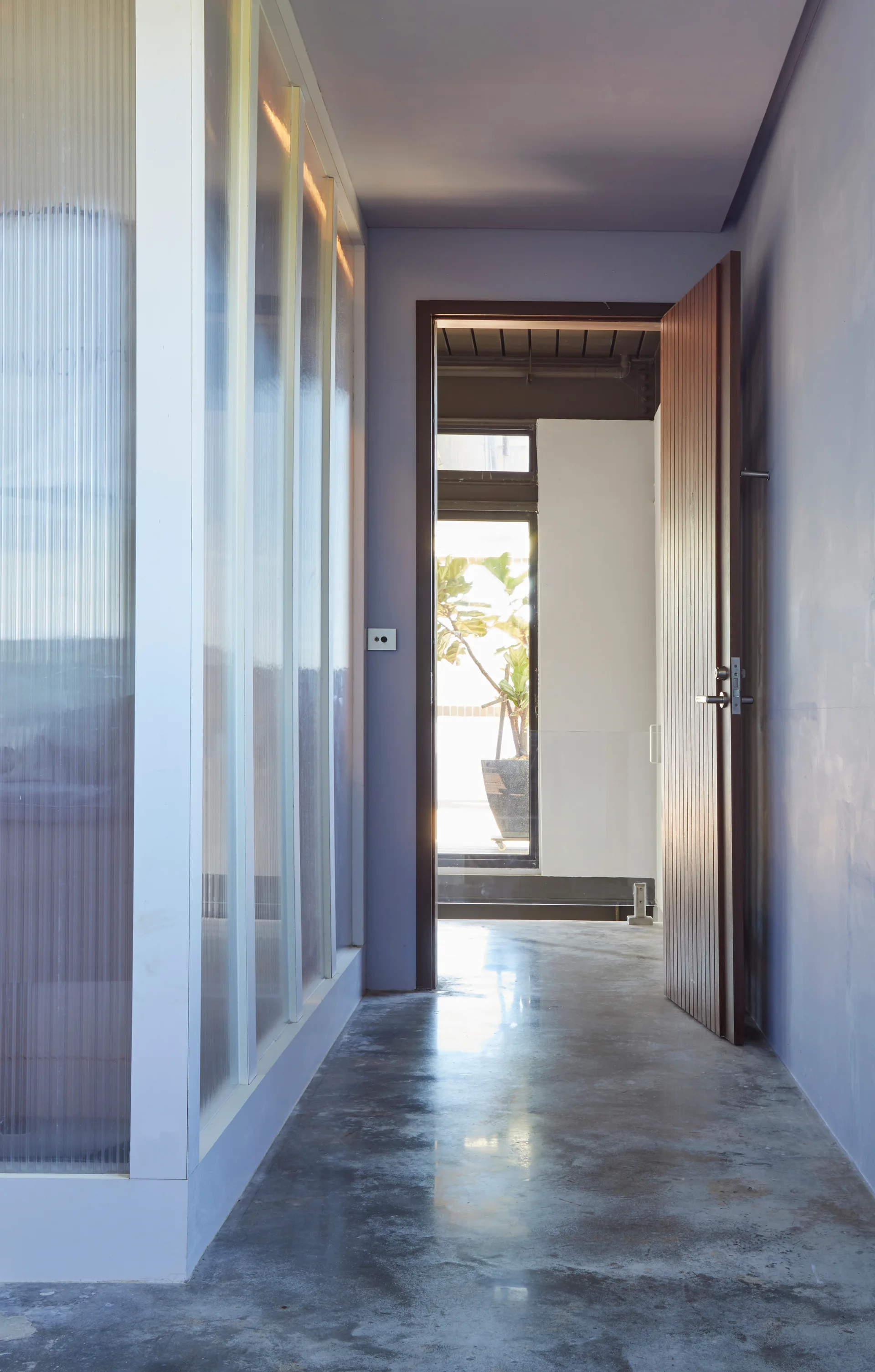 A modern hallway features a polished concrete floor, a translucent ribbed glass partition wall, periwinkle blue walls, and an open slatted wooden door.