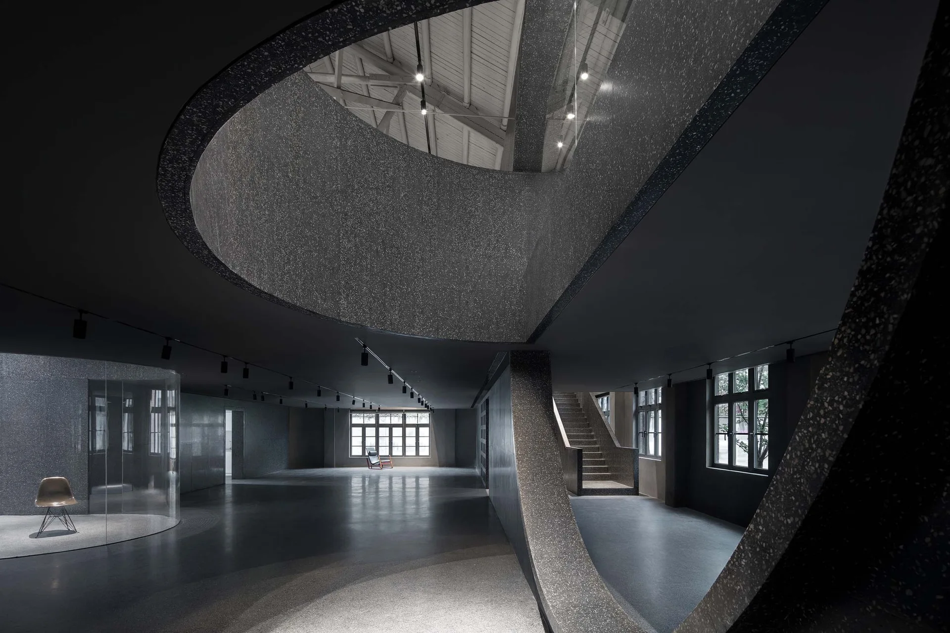 A large minimalist room with a curved charcoal terrazzo staircase, polished concrete floors, and track lighting beneath an open ceiling structure.