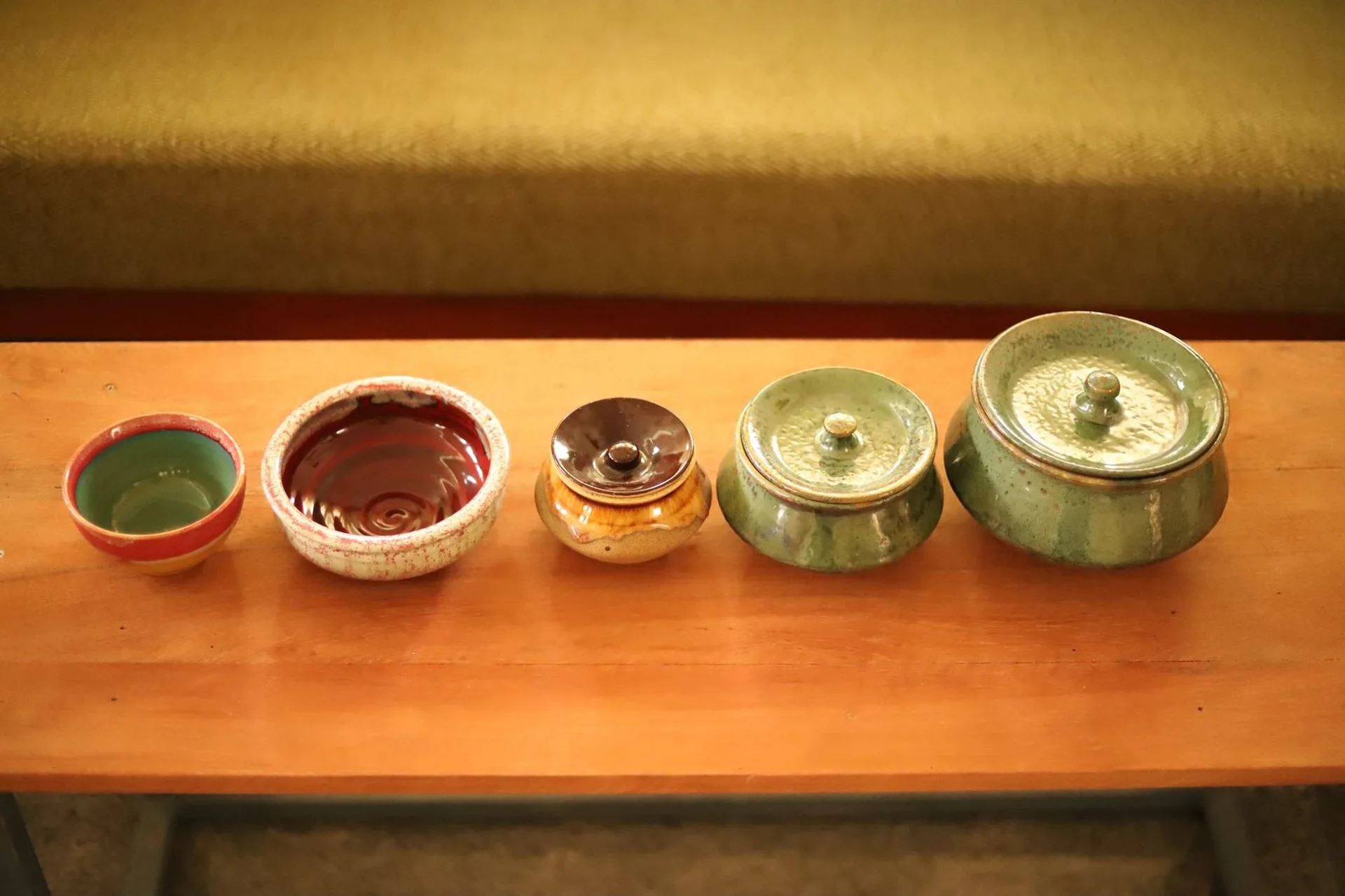 Five glazed ceramic bowls and lidded jars in rich earth tones are displayed in a neat row on a wooden tabletop.