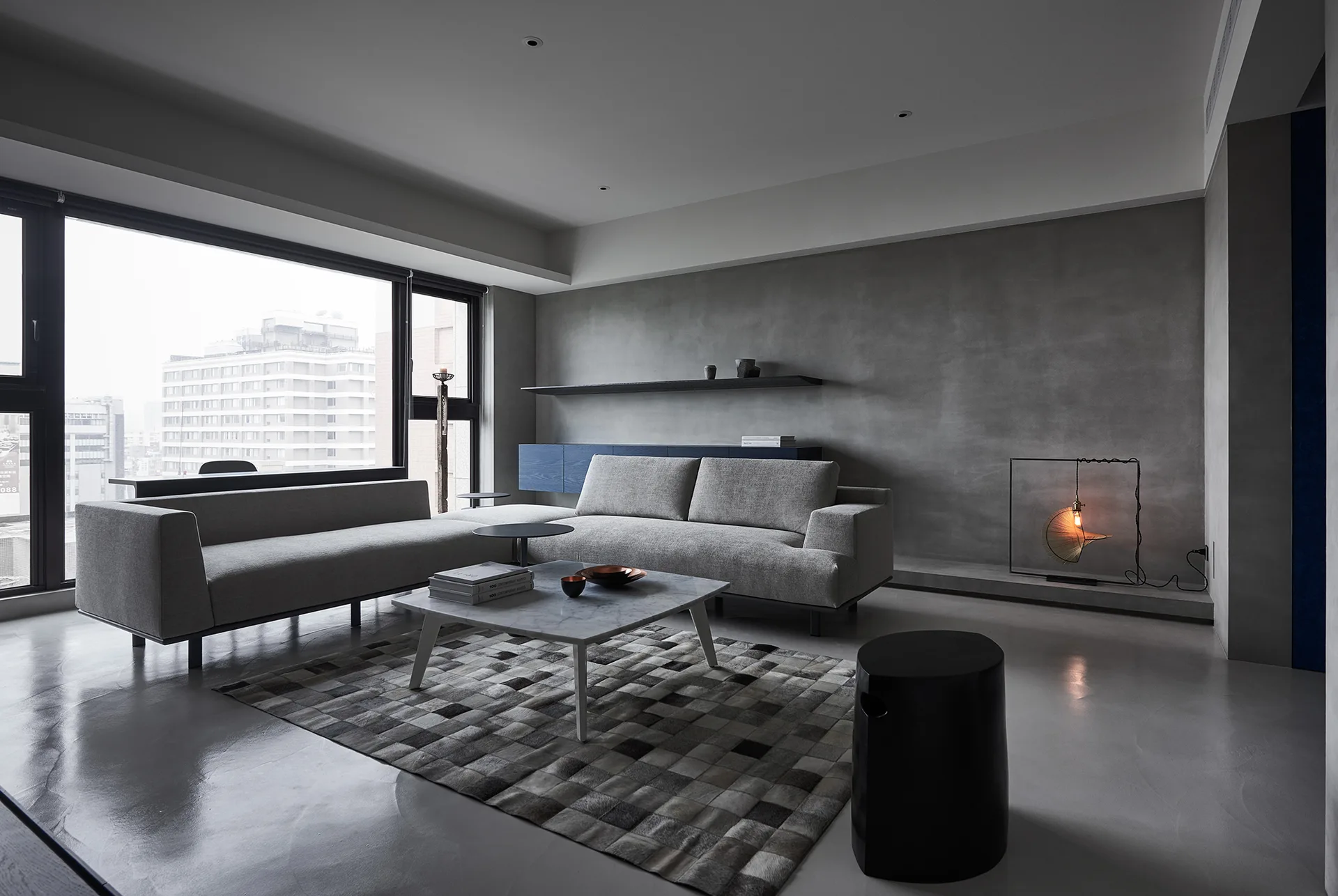 A minimalist living room features a grey sectional sofa, marble-top coffee table, and a checkered rug against textured grey concrete walls.