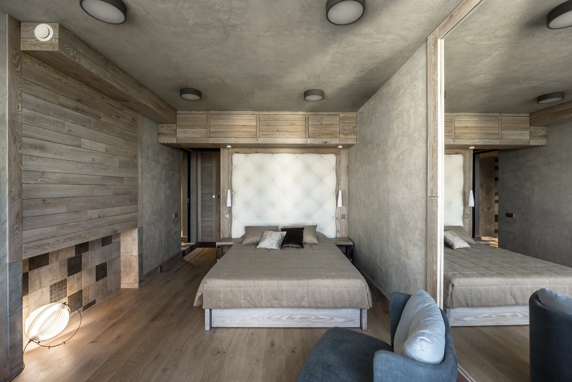 A contemporary bedroom with a white tufted headboard under wood cabinets, plaster walls, and a large mirror reflecting the bed.