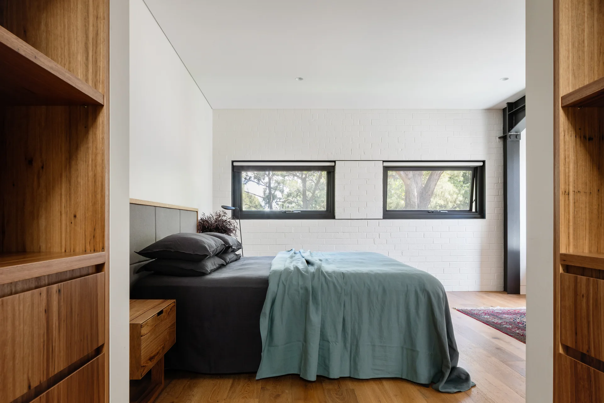 A modern industrial bedroom featuring a bed with charcoal and teal linens set against a white painted brick wall with two black-framed windows.