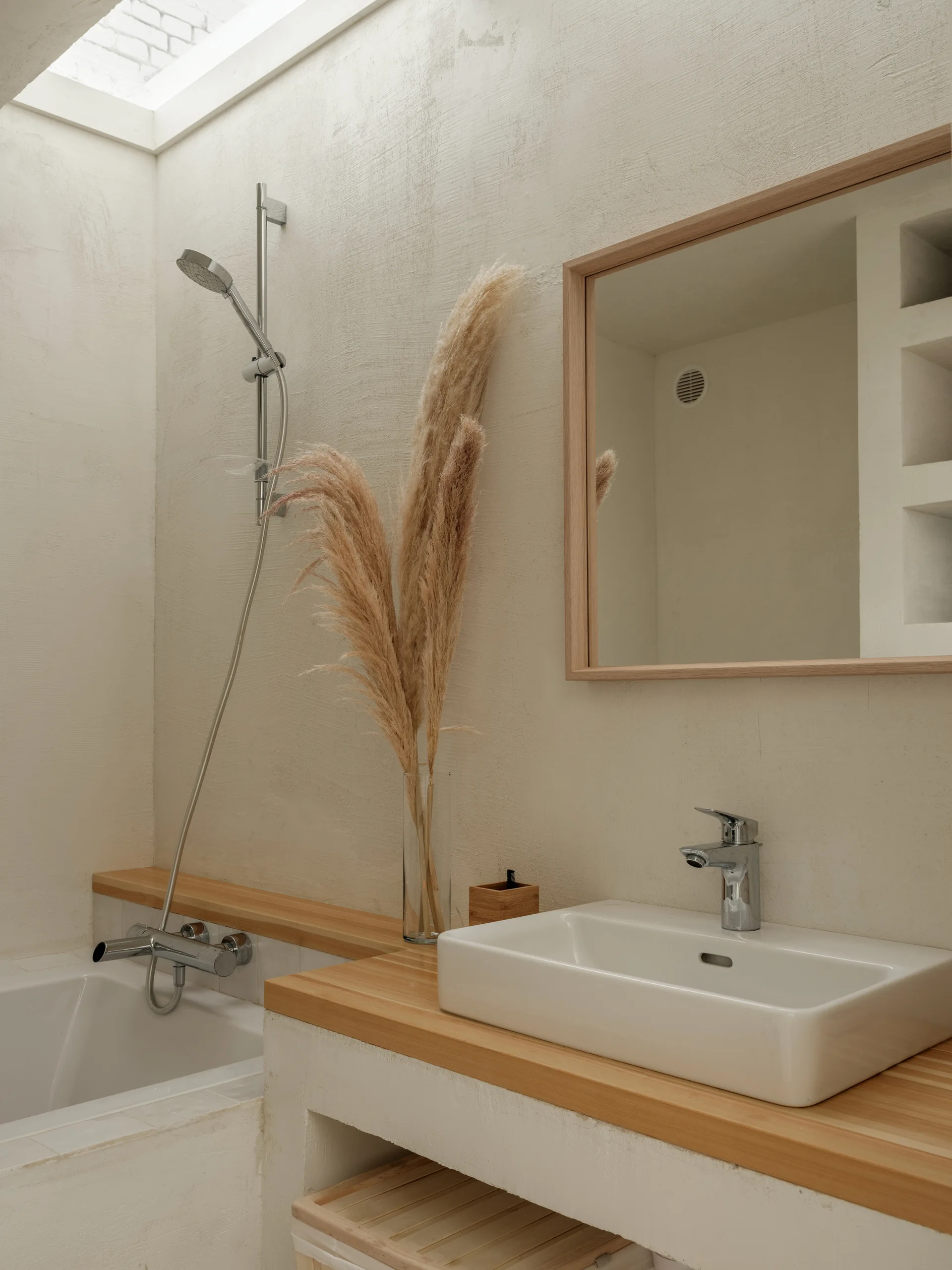 A minimalist bathroom vanity featuring a wooden countertop, white ceramic sink, wood-framed mirror, and textured cream plaster walls under a skylight.