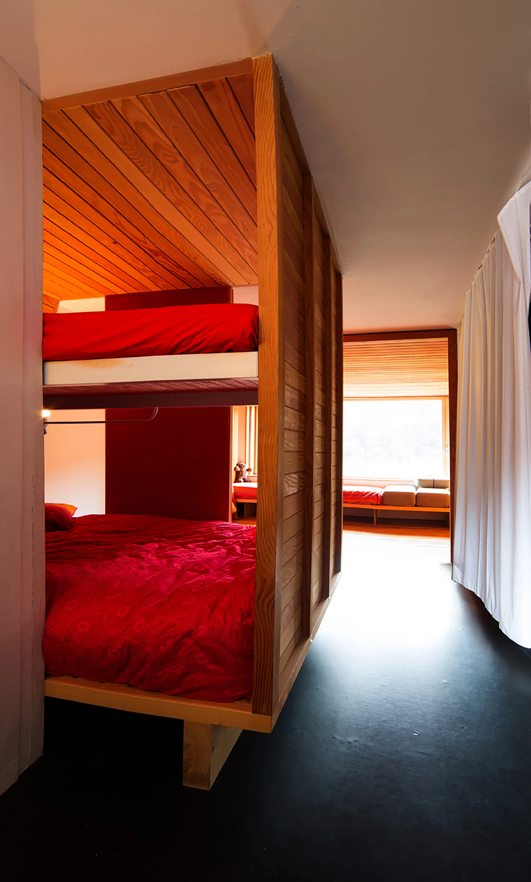 A modern bedroom featuring a built-in wooden bunk bed with red bedding, wood-paneled walls, and a slatted ceiling.