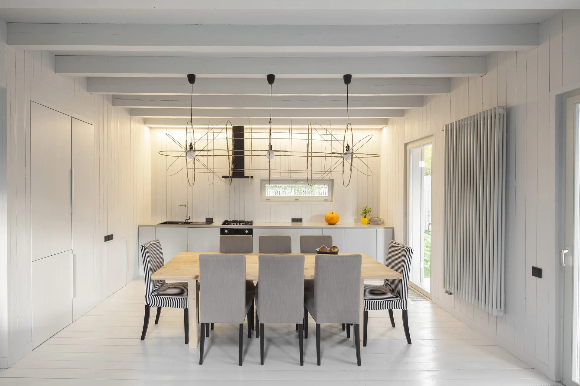 A bright dining room featuring a light wood table, grey and striped chairs, white vertical wall paneling, and large wire pendant lights.