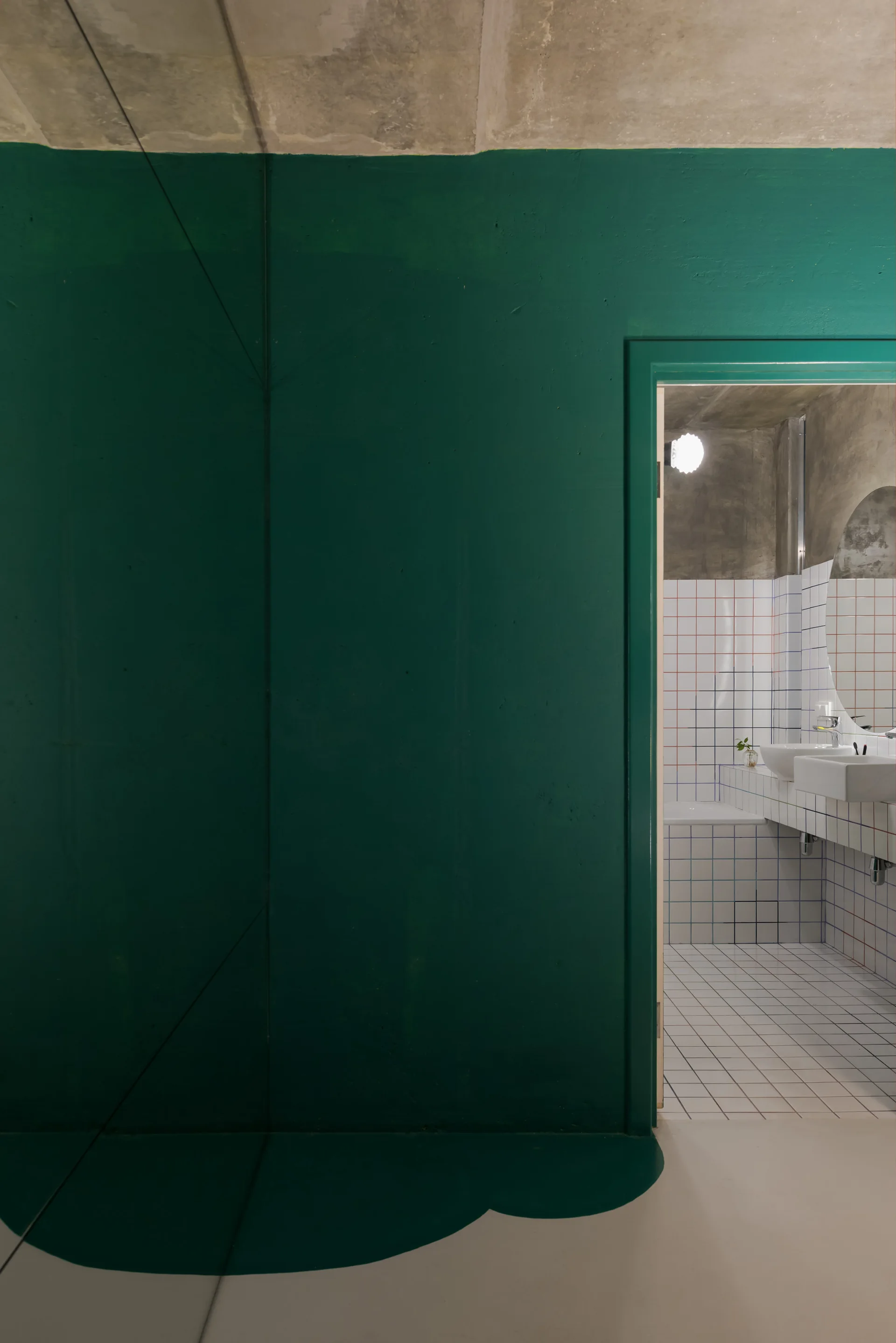 A view through a doorway in a dark green wall reveals a bathroom finished with white grid tiles, a concrete ceiling, and a white sink.