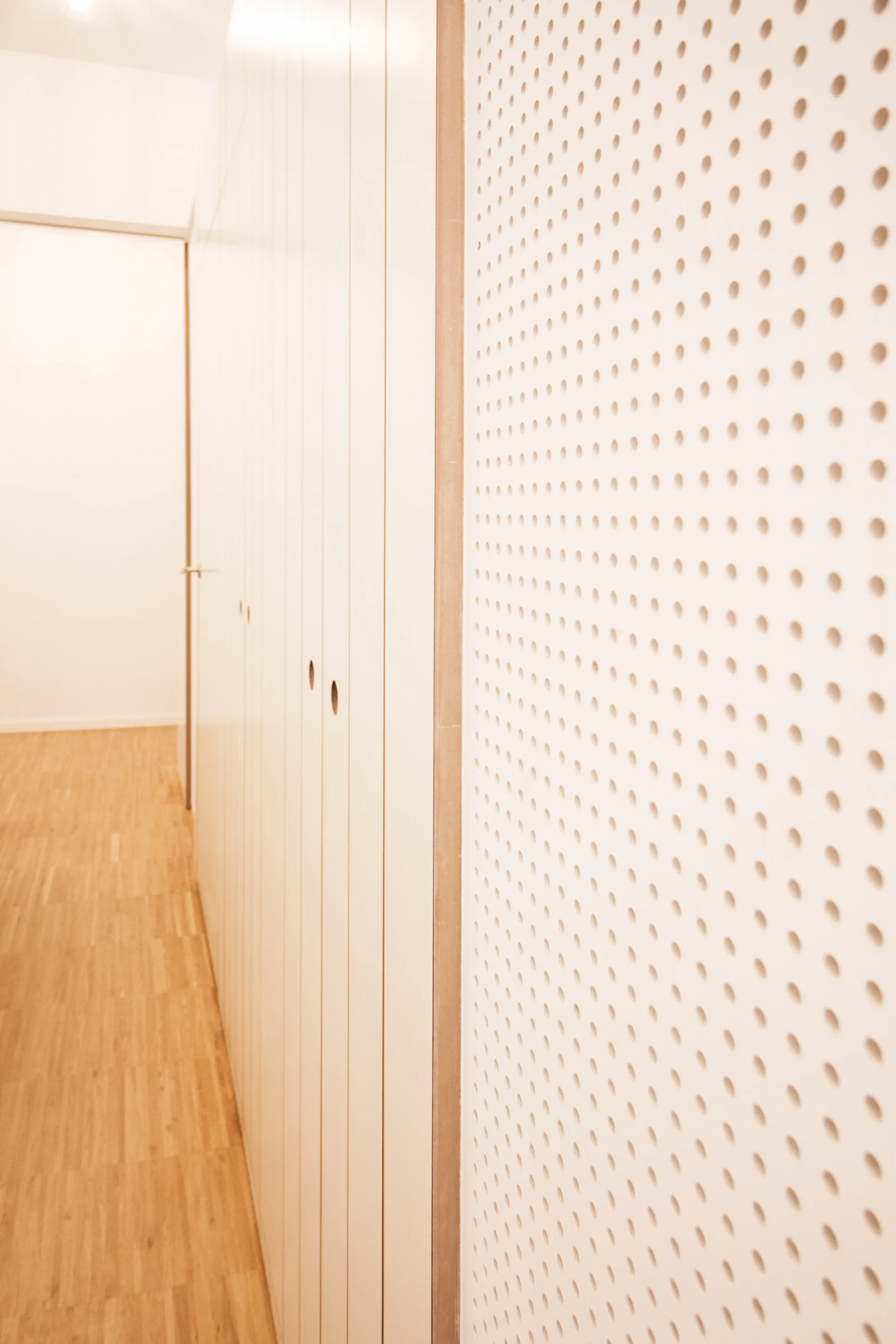 Minimalist hallway featuring off-white built-in cabinets with circular finger pulls, light wood flooring, and a perforated wall panel.