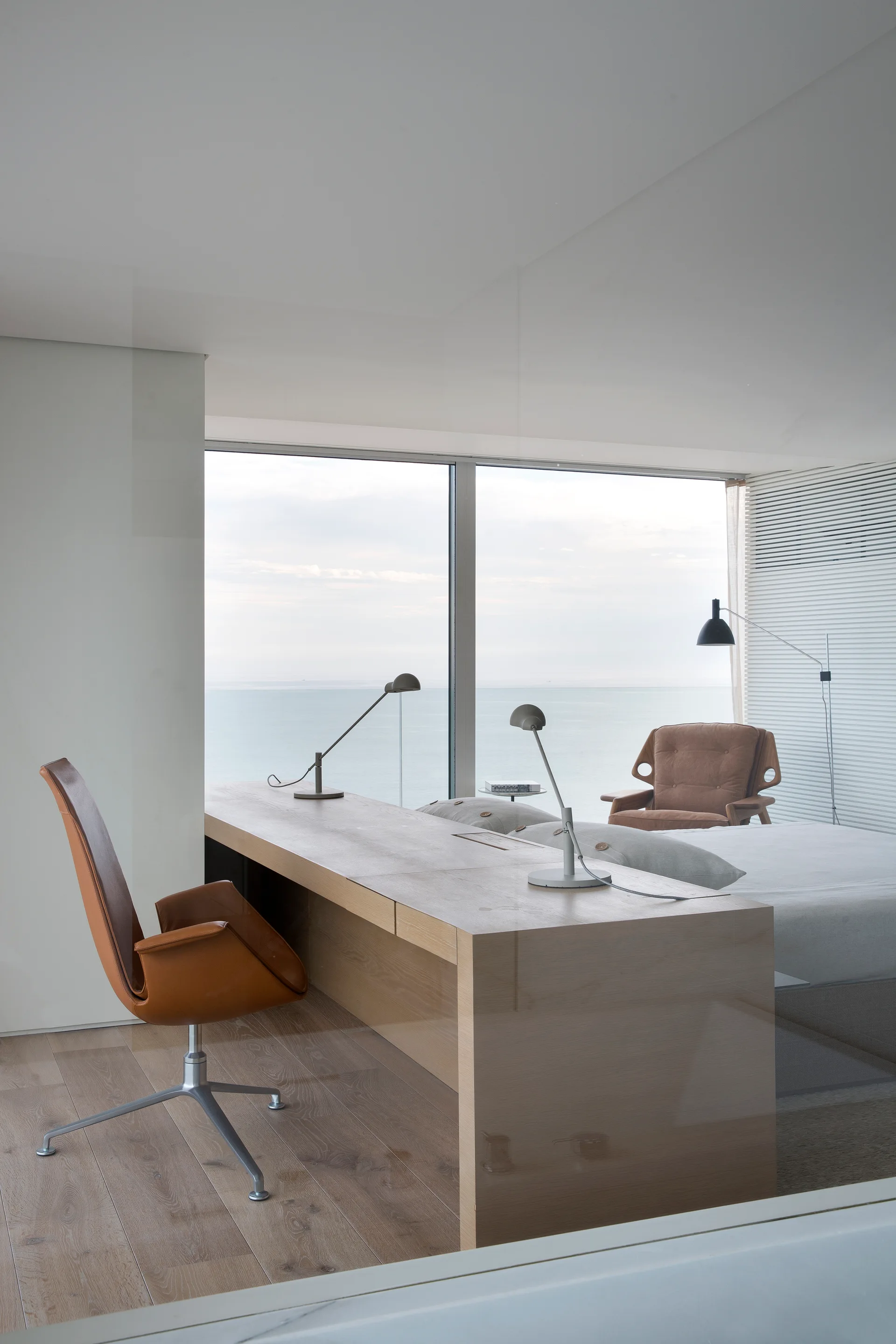 A modern bedroom features a light wood desk, a brown leather swivel chair, and a bed set against floor-to-ceiling windows with an ocean view.
