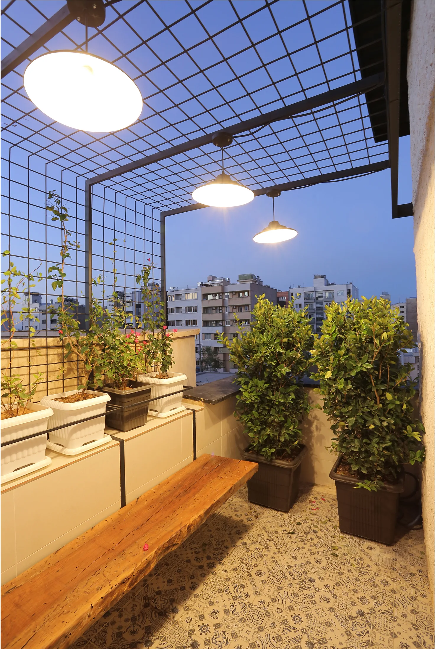 An outdoor terrace at twilight with a live-edge wooden bench, potted green plants, and warm pendant lights suspended from a black metal grid.