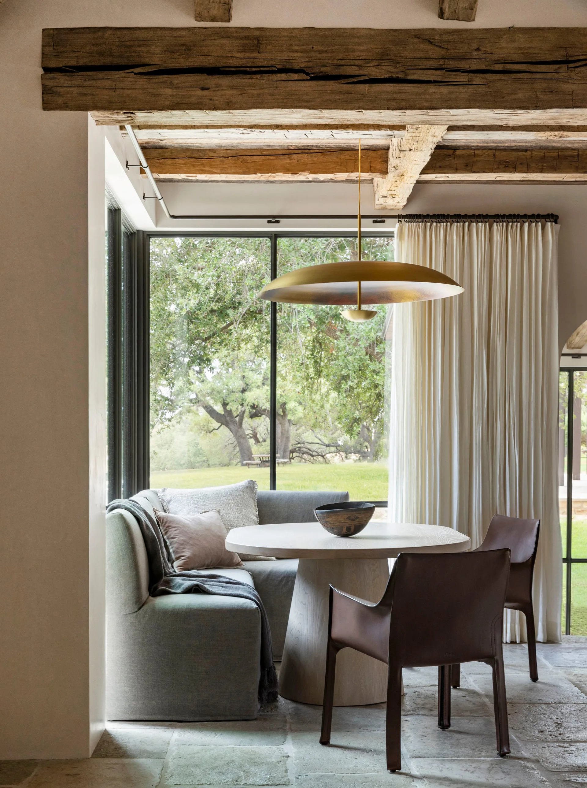 A breakfast nook features a circular wooden table, a grey upholstered banquette, and brown leather chairs beneath exposed timber ceiling beams.