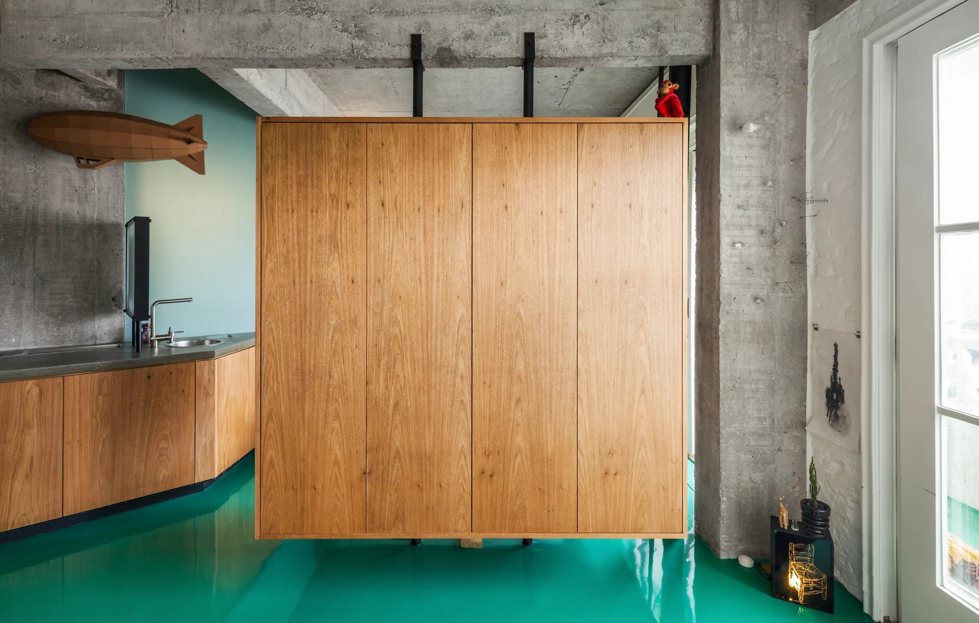 An industrial loft kitchen featuring a suspended wooden cabinet, exposed concrete walls, and a high-gloss turquoise epoxy floor.