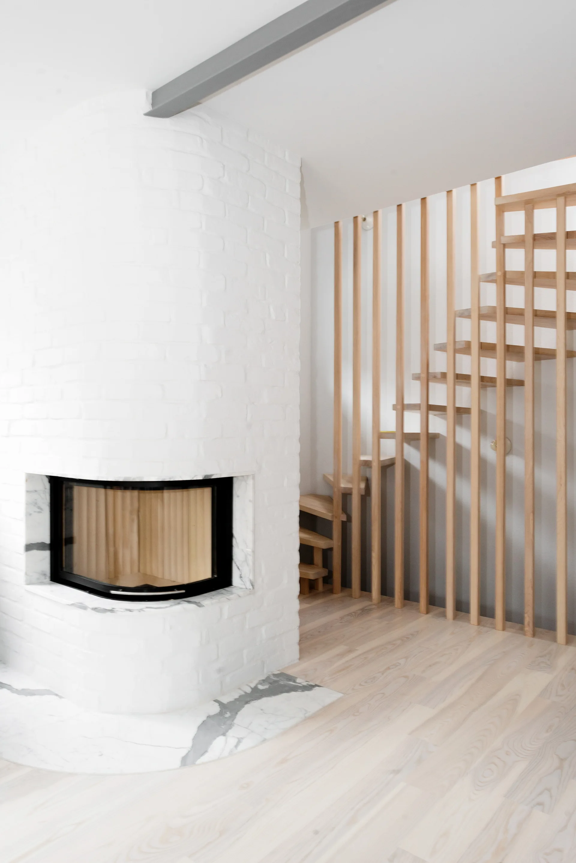 A modern room featuring a curved white brick fireplace with a marble hearth and a wooden staircase with vertical slat railings.