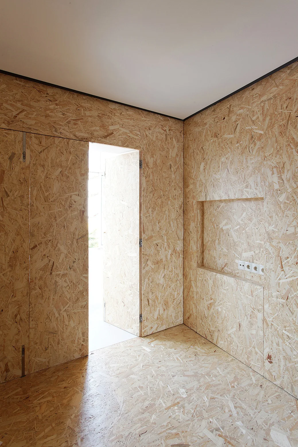 A minimalist room features walls and floors clad in oriented strand board, complete with a recessed wall niche and an open seamless door.