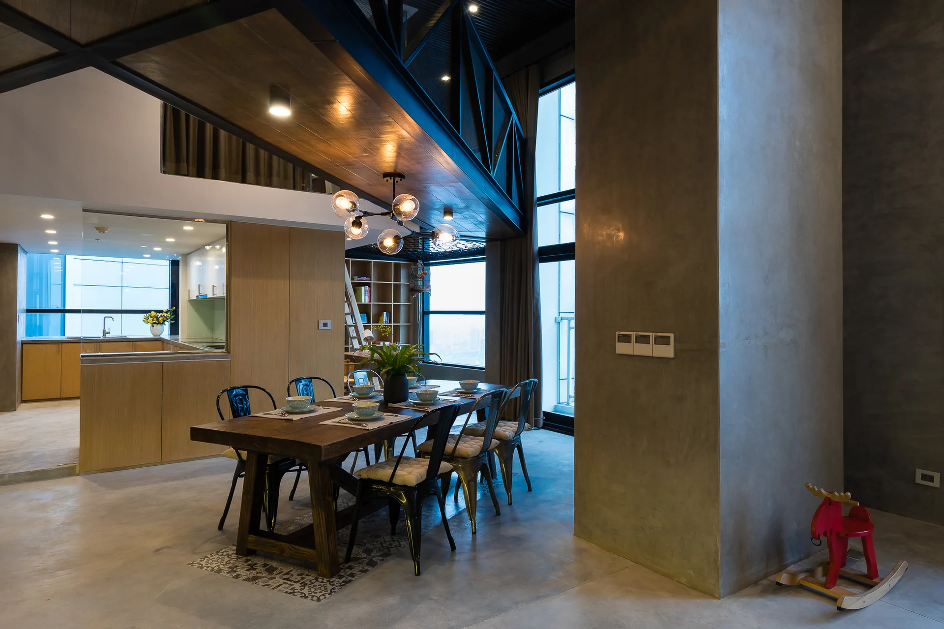 A loft dining room features a wooden dining table, metal chairs, concrete walls, a globe pendant light, and large windows.