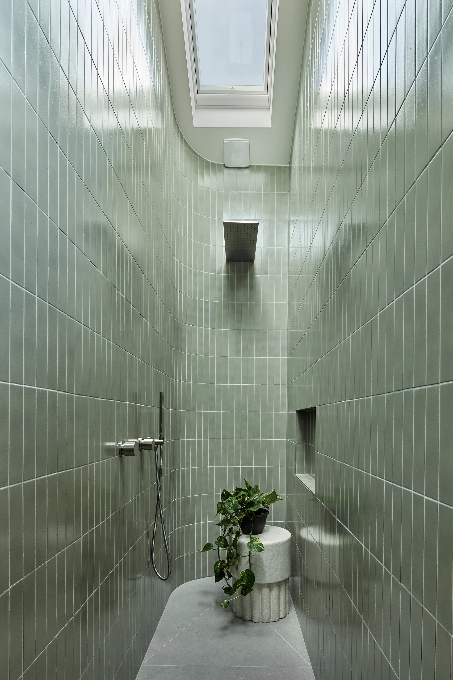 A narrow walk-in shower features vertical sage green wall tiles, a curved wall, a skylight, and a concrete stool with a potted plant.