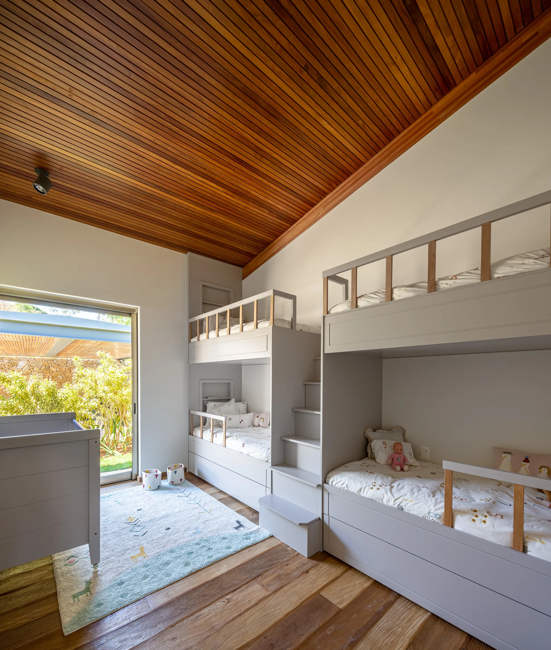 A children's bedroom featuring a custom grey built-in bunk bed unit, a matching crib, and a vaulted wood slat ceiling next to a sliding glass door.