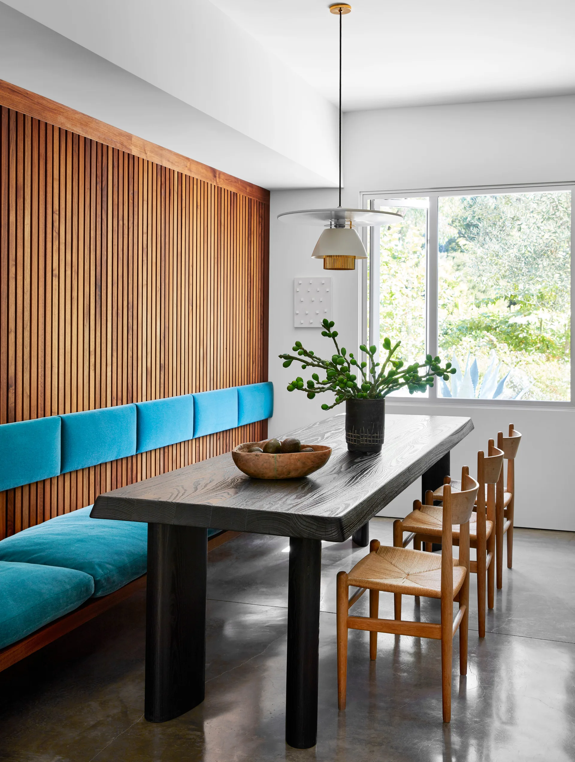 A modern dining room featuring a dark wood table, woven-seat chairs, and a long teal upholstered bench set against a vertical slatted wood wall.