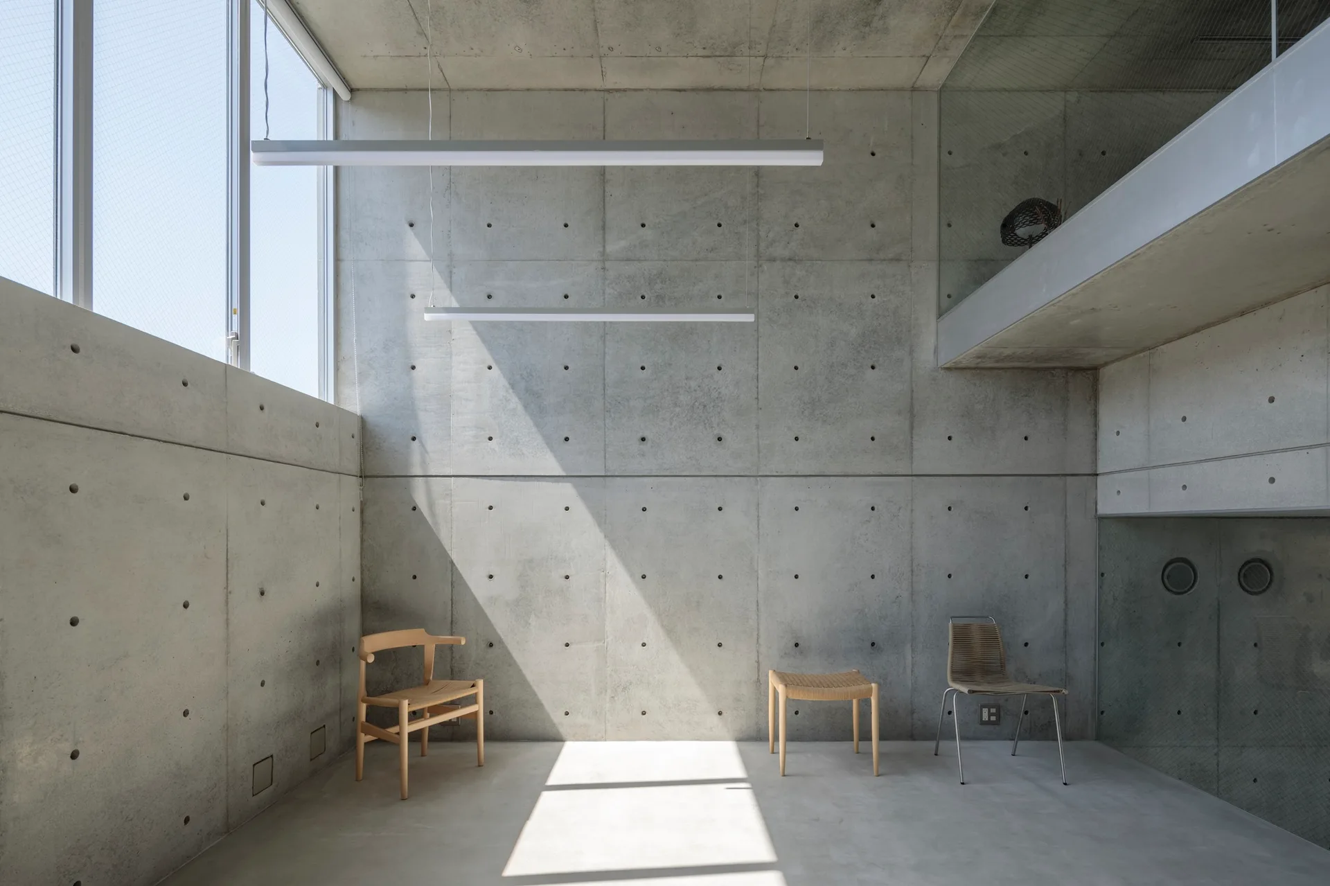 A minimalist room with raw cast concrete walls and floor features three different wooden and metal chairs arranged under hanging linear lights.