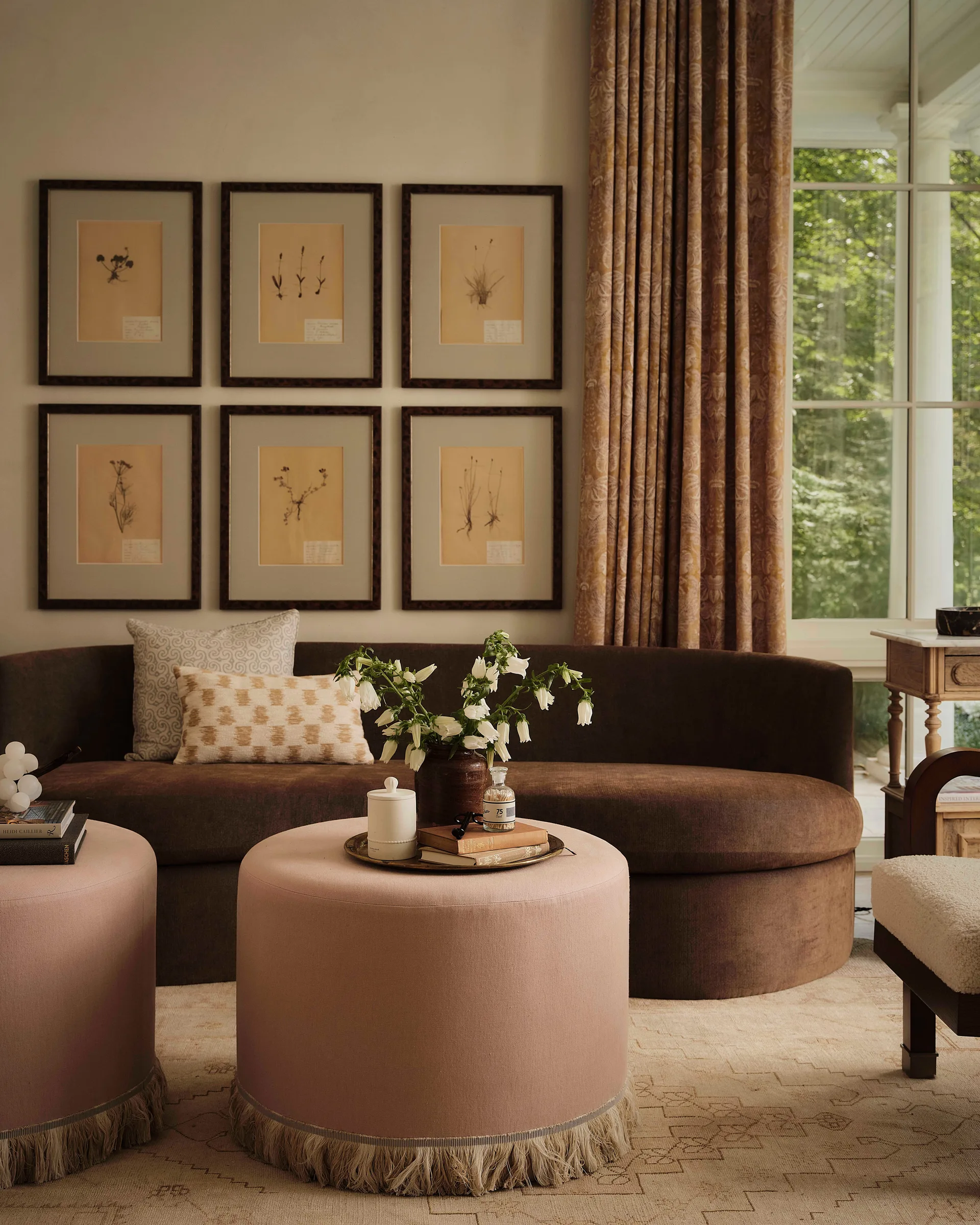 A curved brown sofa sits beneath a grid of six framed botanical prints, paired with two round blush pink ottomans with fringed bases.