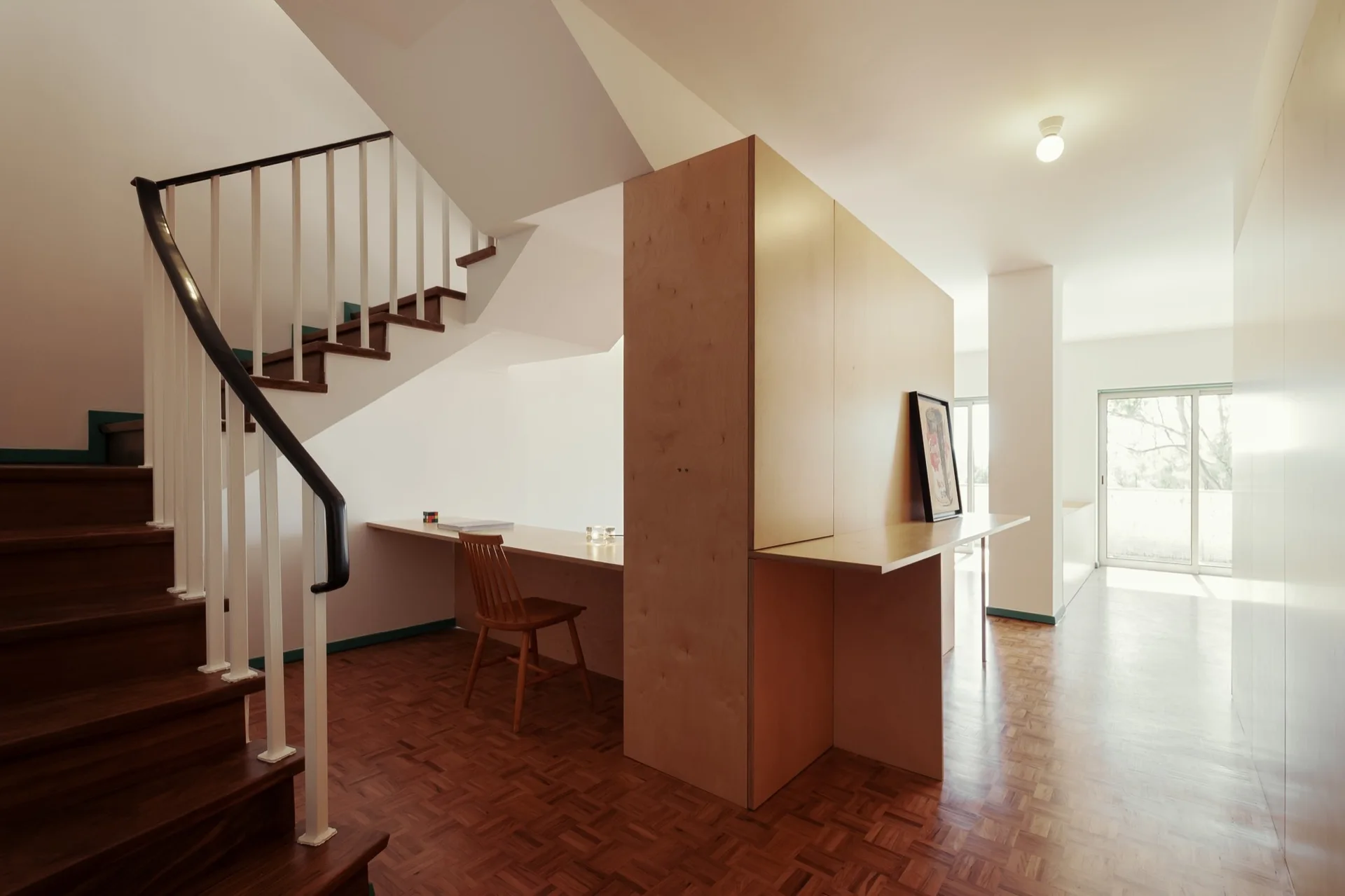 A minimalist interior showing a wood staircase next to a light plywood partition with an integrated writing desk, chair, and parquet flooring.