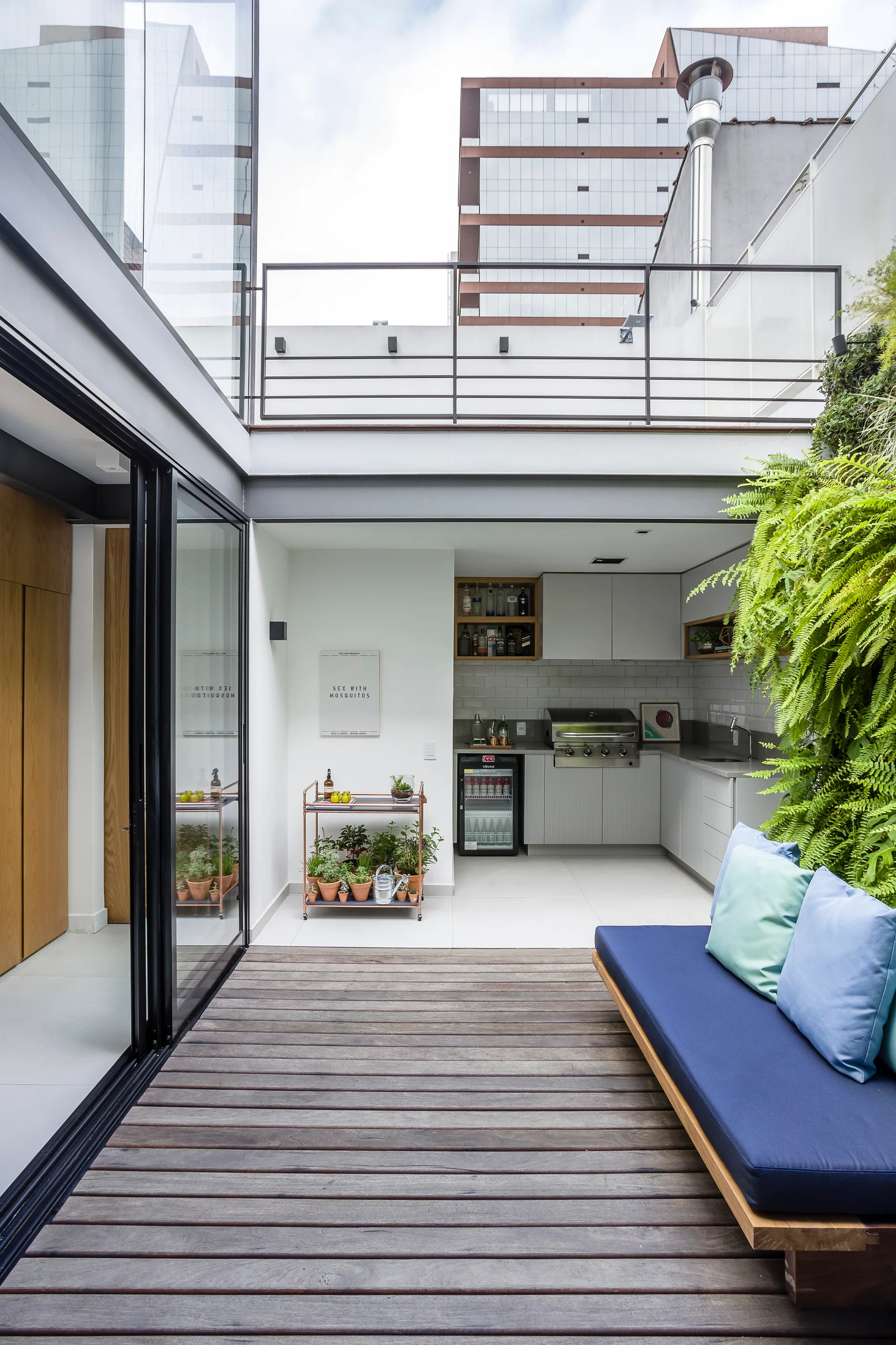 A modern outdoor courtyard featuring wood decking, a blue platform sofa, a vertical green garden, and a built-in grey kitchen.