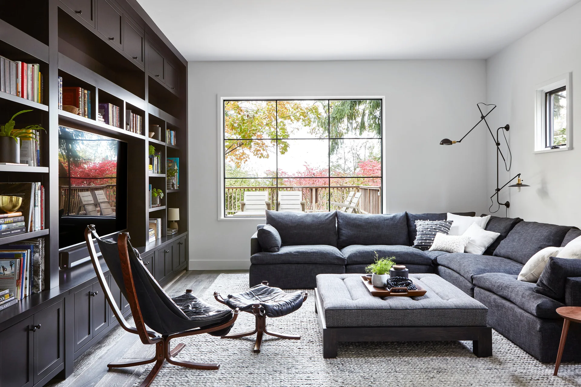 A modern living room features a dark built-in media wall, a charcoal sectional sofa, a textured area rug, and a large black grid window.