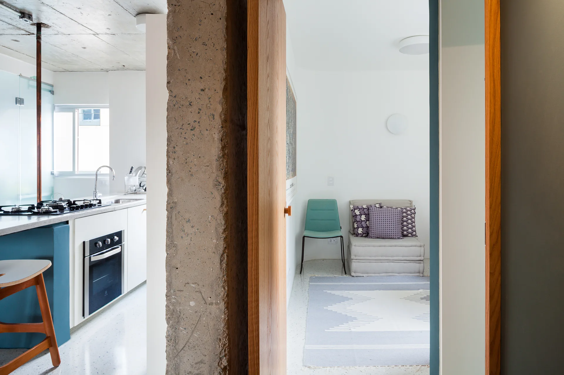 An open wooden doorway next to an exposed concrete pillar reveals a modern kitchen on the left and a small sitting area with a rug on the right.