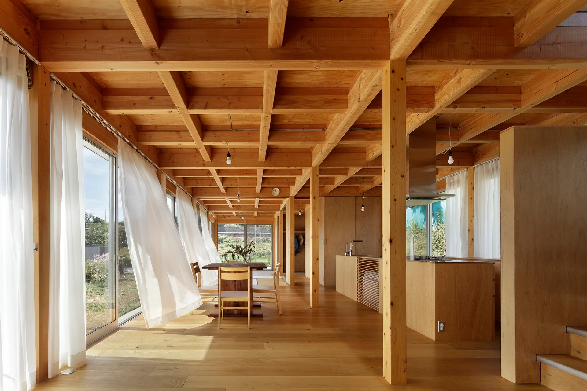 An open-plan room with exposed timber post and beam framing, a wooden dining set, a wood kitchen island, and sheer white curtains.