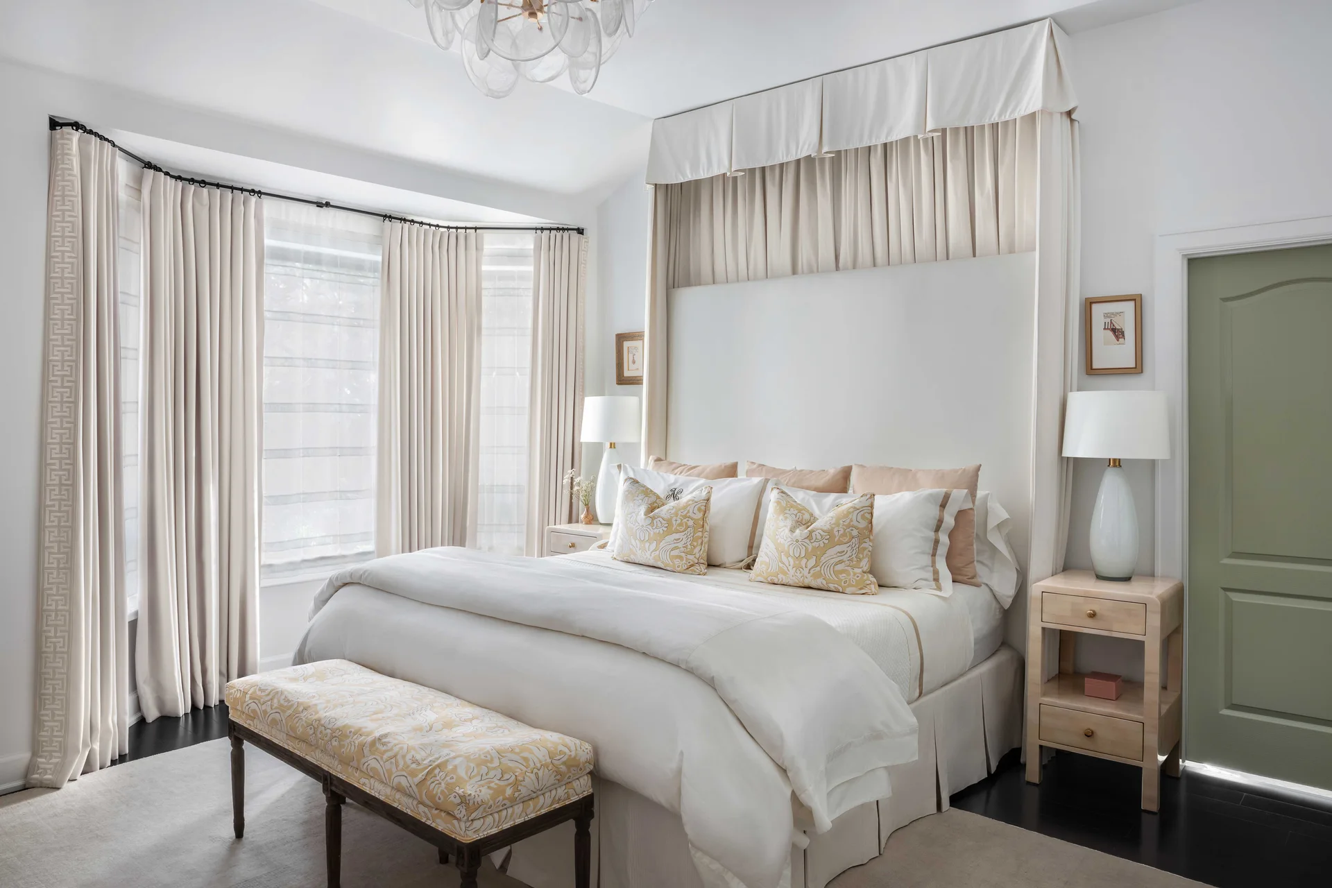 A well-lit bedroom features a tall white canopy bed, cream-colored drapery on a bay window, and a sage green painted door.