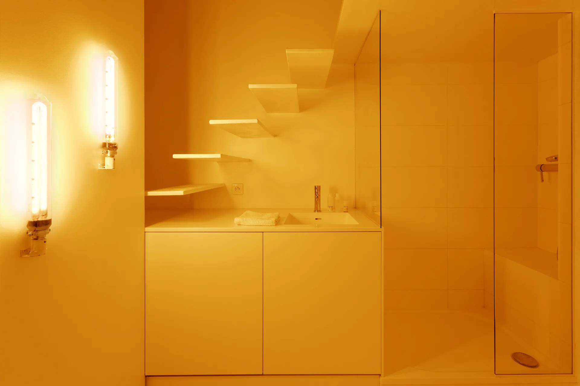 A minimalist white bathroom featuring a modern vanity, staggered floating shelves, a glass-enclosed shower, and glowing tube sconces.