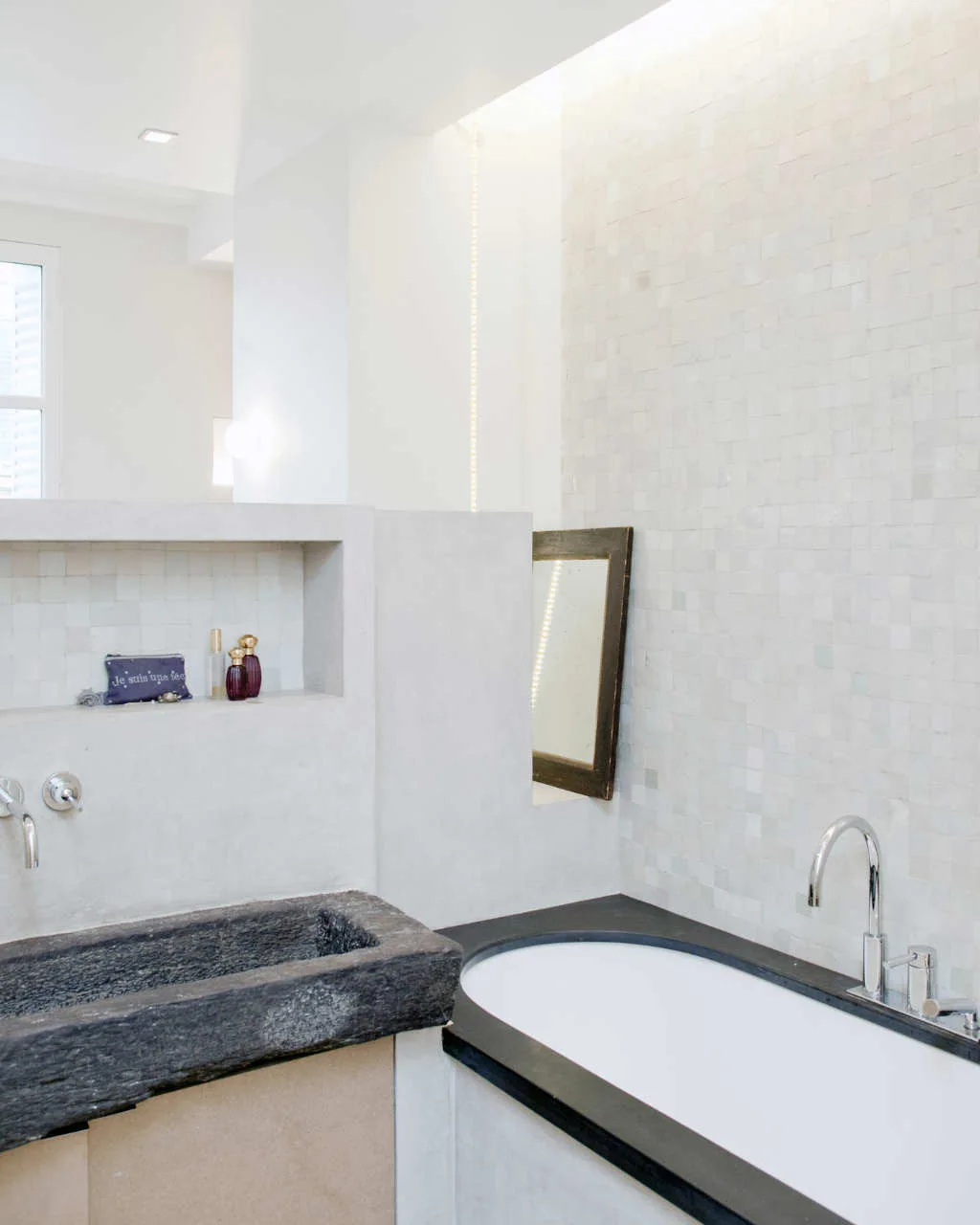 A contemporary bathroom featuring a rustic stone sink and a white bathtub separated by a grey concrete partition with a built-in niche.