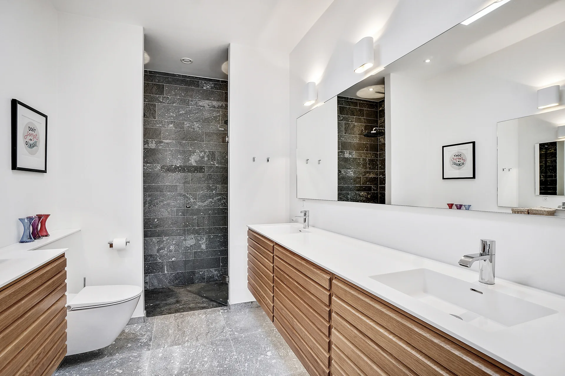 A modern bathroom featuring a horizontal slatted wood double vanity, a wide wall mirror, and a walk-in shower lined with dark grey stone tiles.