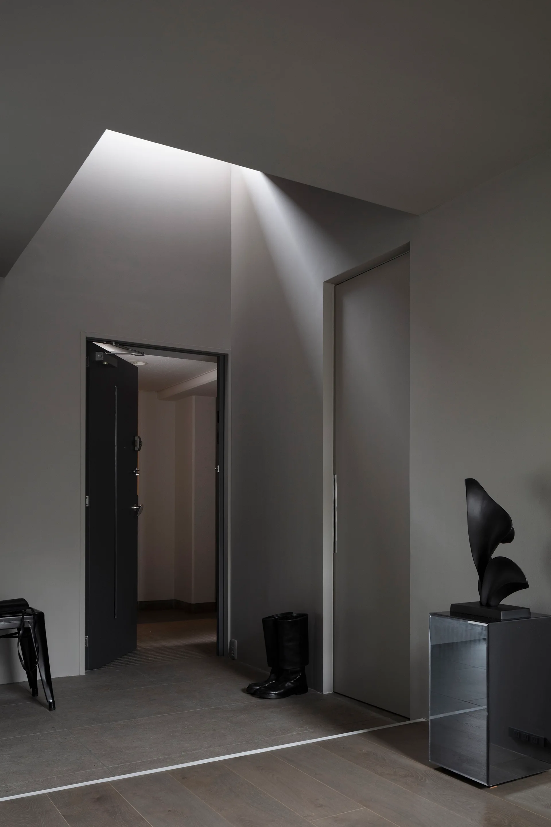 A minimalist grey entryway with a skylight casting light near a mirrored pedestal holding an abstract black sculpture.