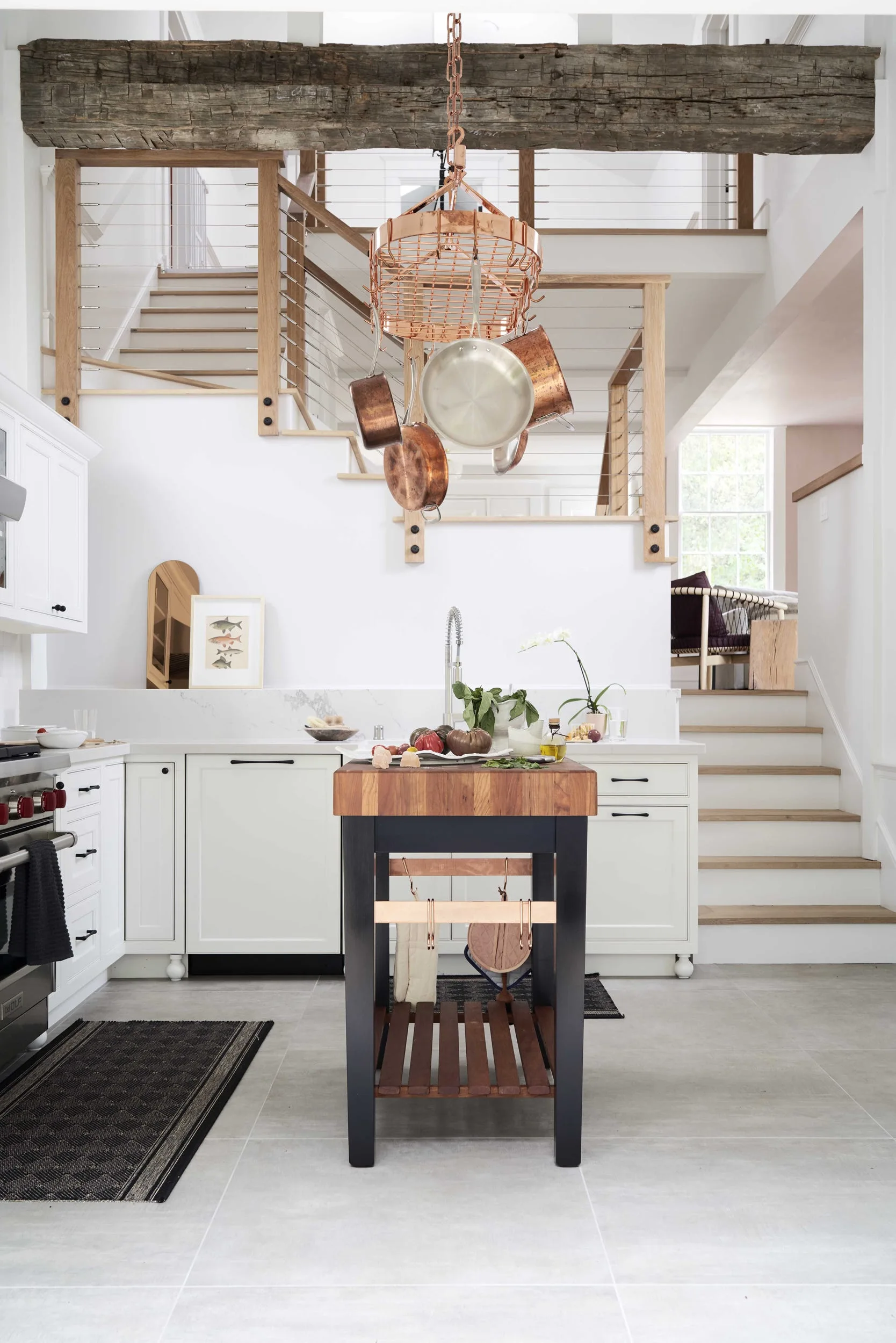 A light-filled kitchen featuring a black island with a butcher block top beneath a hanging copper pot rack and a rustic wooden beam.