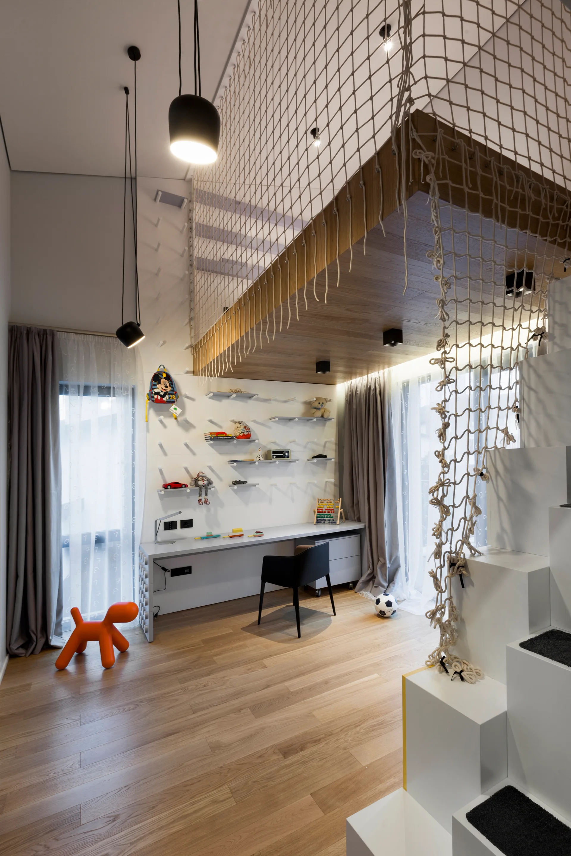 A modern children's bedroom features a wood-toned loft bed with safety netting above a study desk and a white block staircase.