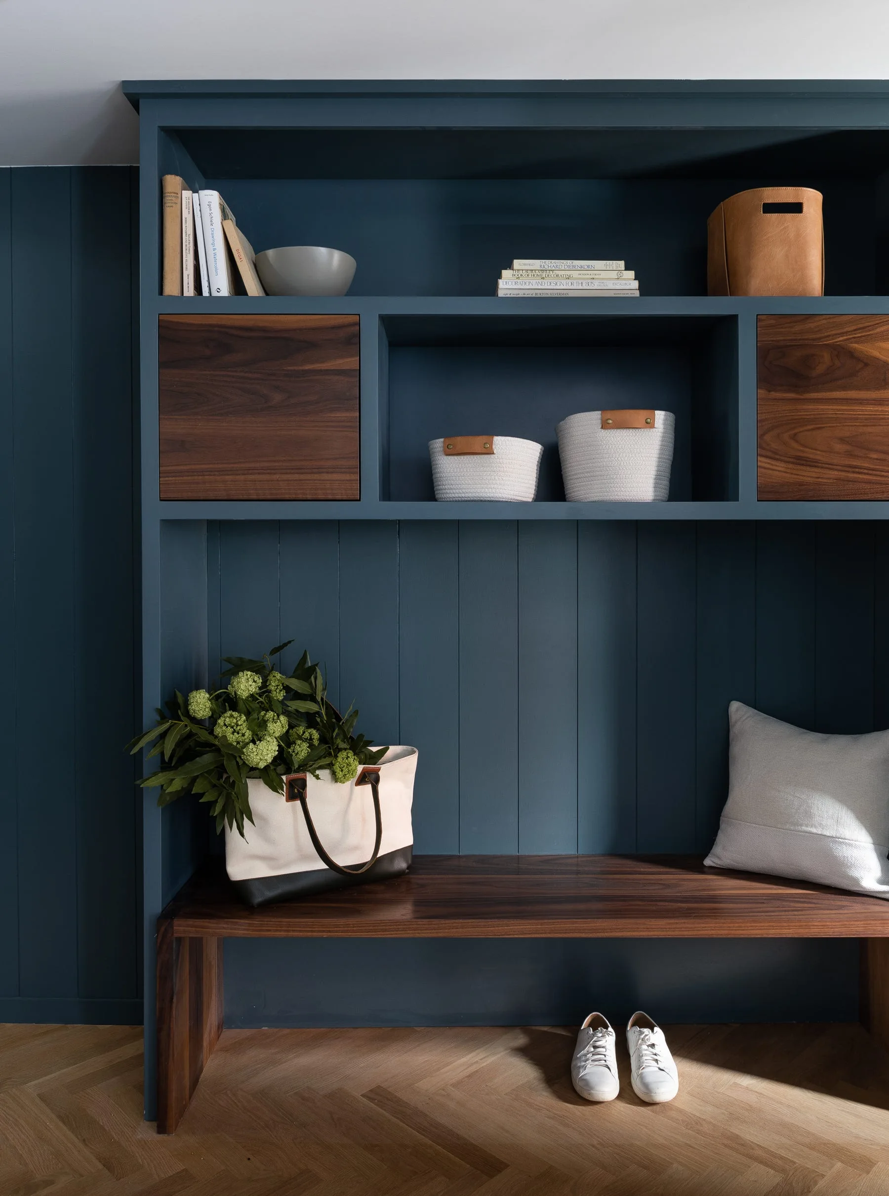 A custom navy blue built-in entryway unit features a dark wood bench, open shelving with decorative baskets, and light herringbone wood floors.