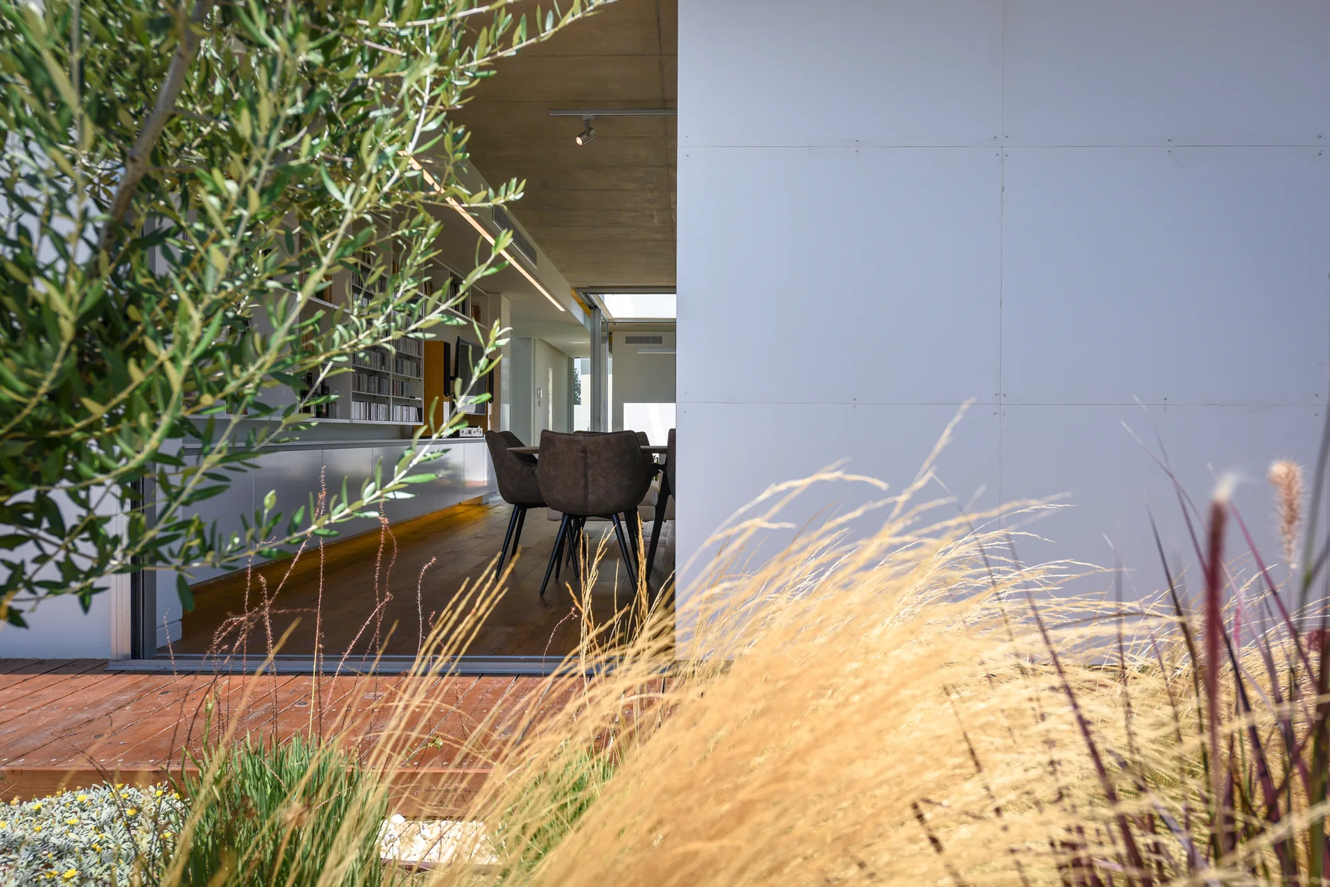 A view from a wooden outdoor deck with tall grasses looking into a modern dining room with dark upholstered chairs and a concrete ceiling.