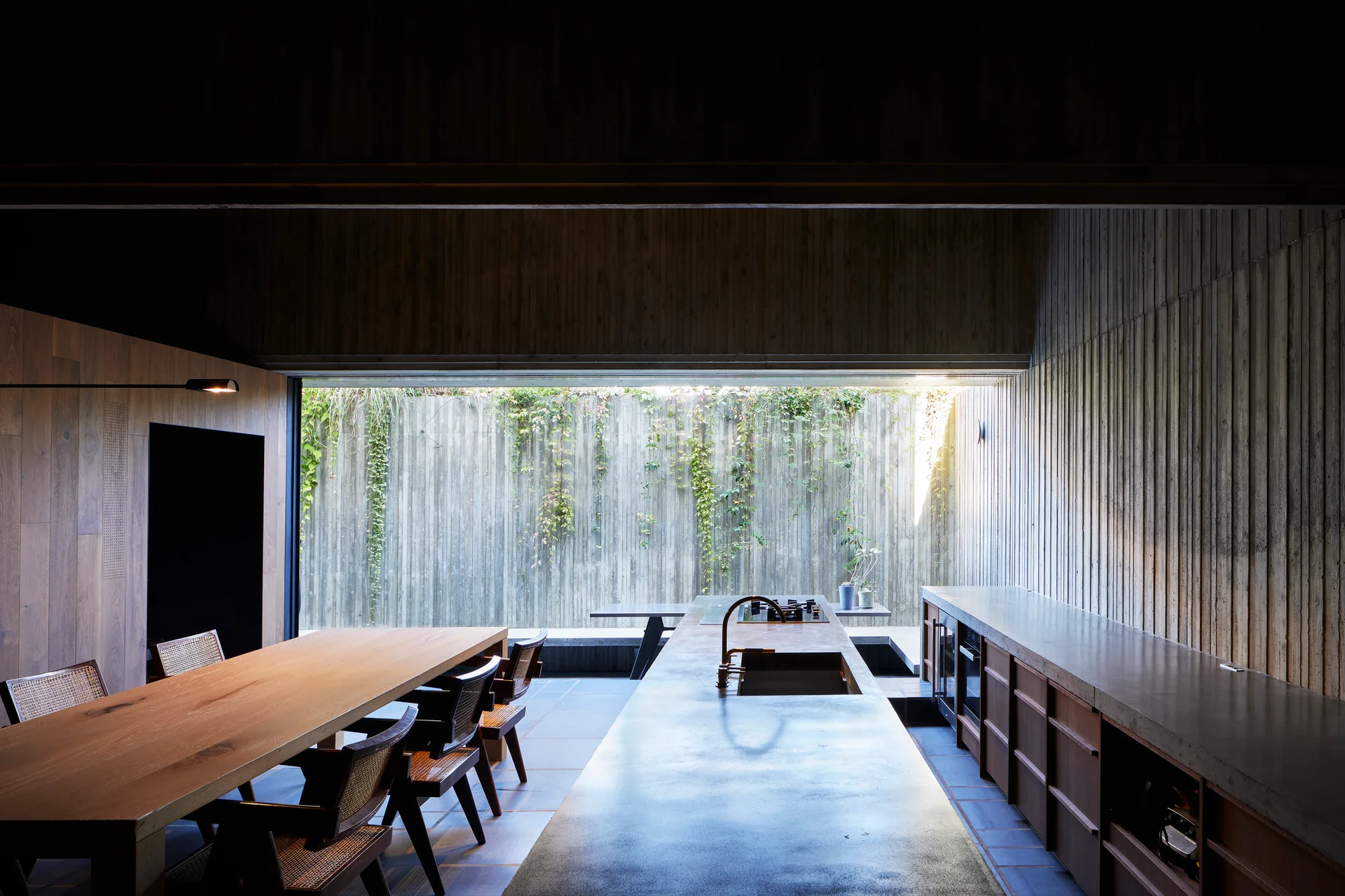 Minimalist kitchen features a long concrete island, wooden dining table with cane chairs, and a wide horizontal window framing an outdoor ivy wall.