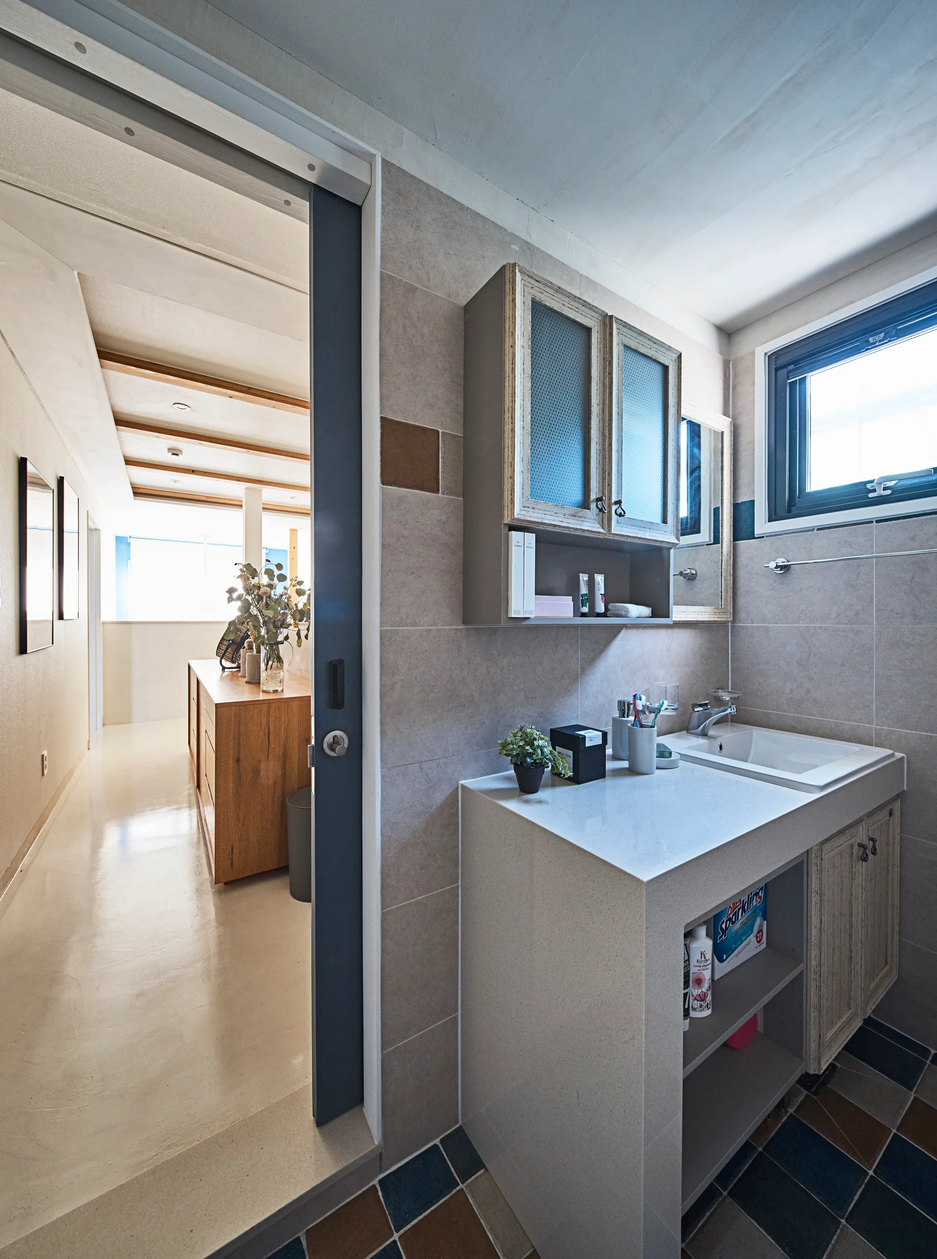 A bathroom features a stone vanity with integrated sink, beige wall tiles, a glass-door cabinet, and a dark sliding door opening to a hallway.